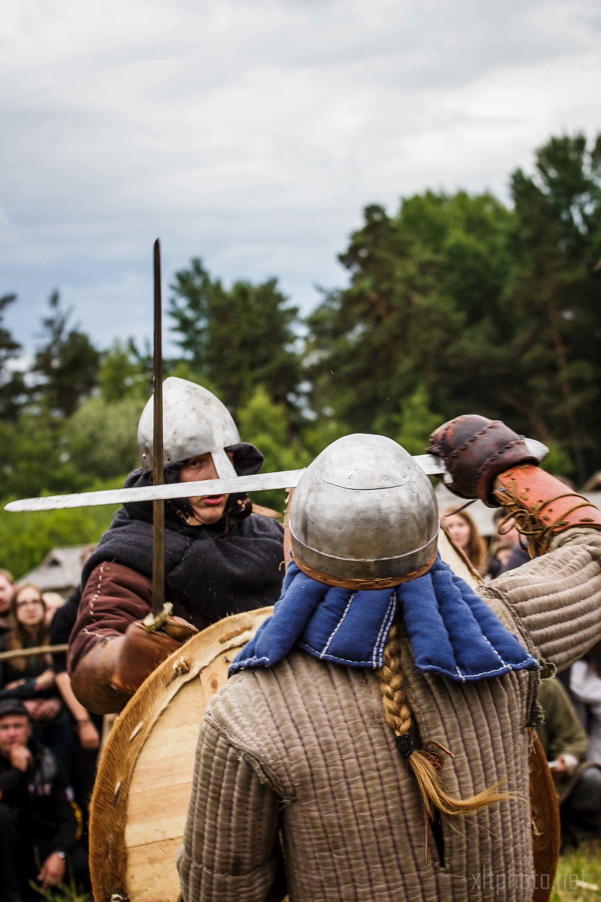 Two people in medieval combat attire are engaged in a sword fight during the Kilkim Žaibu 2015 reenactment event. Both wear helmets and padded clothing. One carries a large round wooden shield, while the other wields a raised sword. A crowd of onlookers and trees are visible in the background.