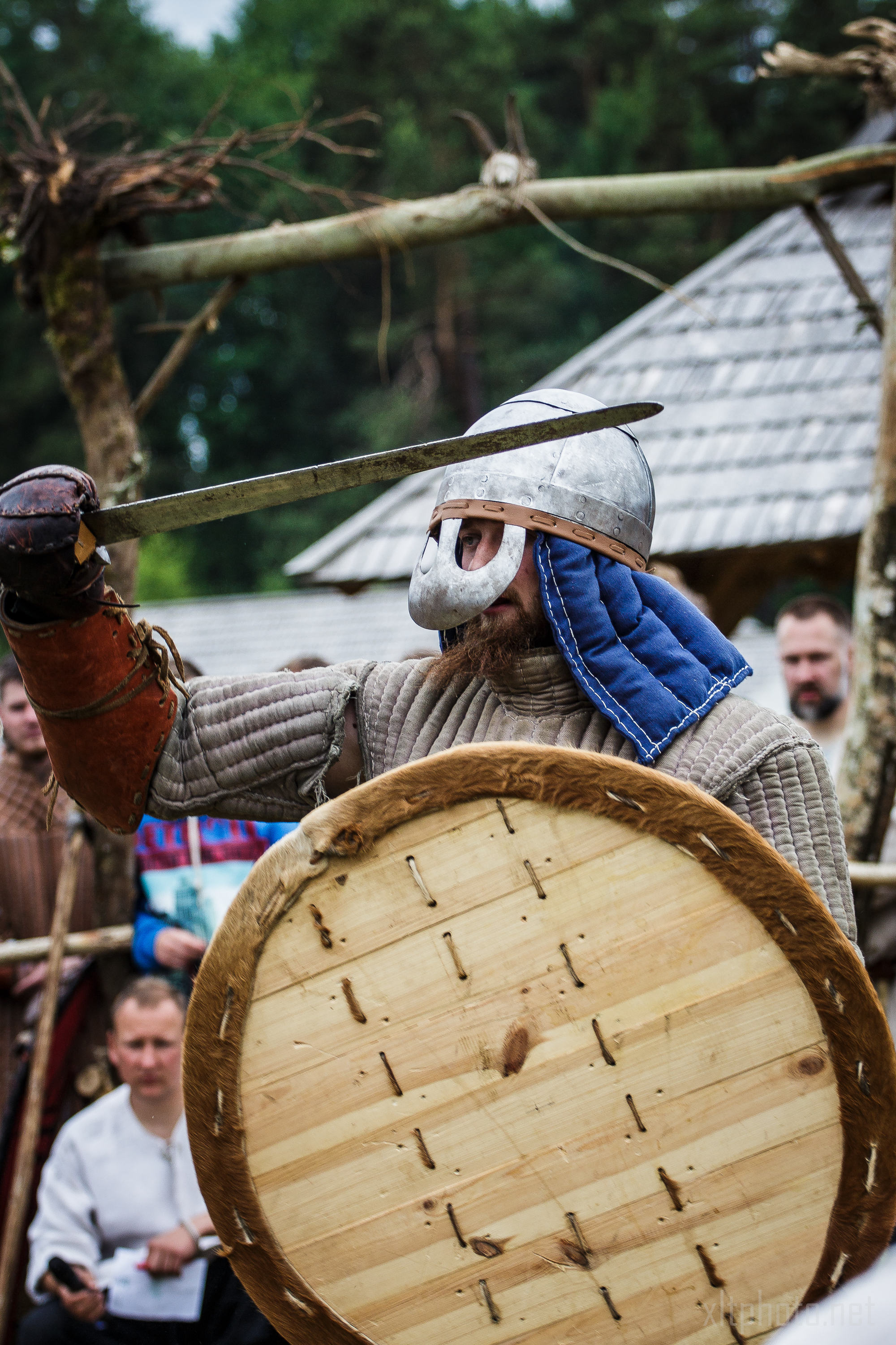 A person dressed in historical warrior attire participates in a reenactment at Kilkim Žaibu 2015. They wear a metal helmet with a blue fabric drape, chainmail, and a padded coat. Holding a round wooden shield in one hand and a drawn sword in the other, they perform as spectators watch the event unfold in the background.