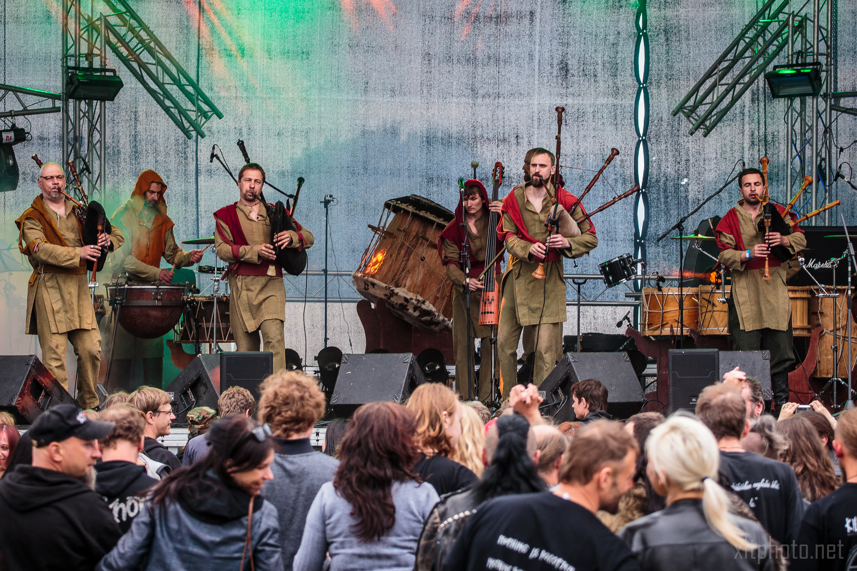 At Kilkim Žaibu 2015, a band performs on a stage at an outdoor event. The group features five musicians playing bagpipes alongside a drummer. Clad in medieval-inspired clothing in brown and red tones, they captivate the crowd gathered before the stage, illuminated by vibrant lighting and speakers.