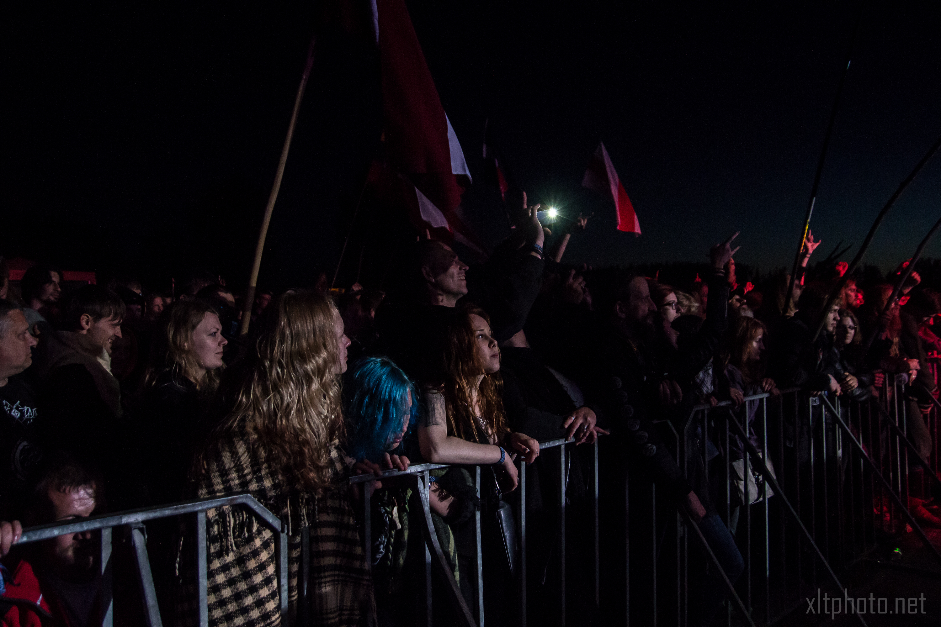A crowd of people at an outdoor festival stands behind a metal barricade, looking towards the stage with intensity. Some hold flags, while others lean forward, engrossed in the Kilkim Žaibu performance. The image is dimly lit, reflecting a nighttime setting with a spotlight casting a red hue on some faces.