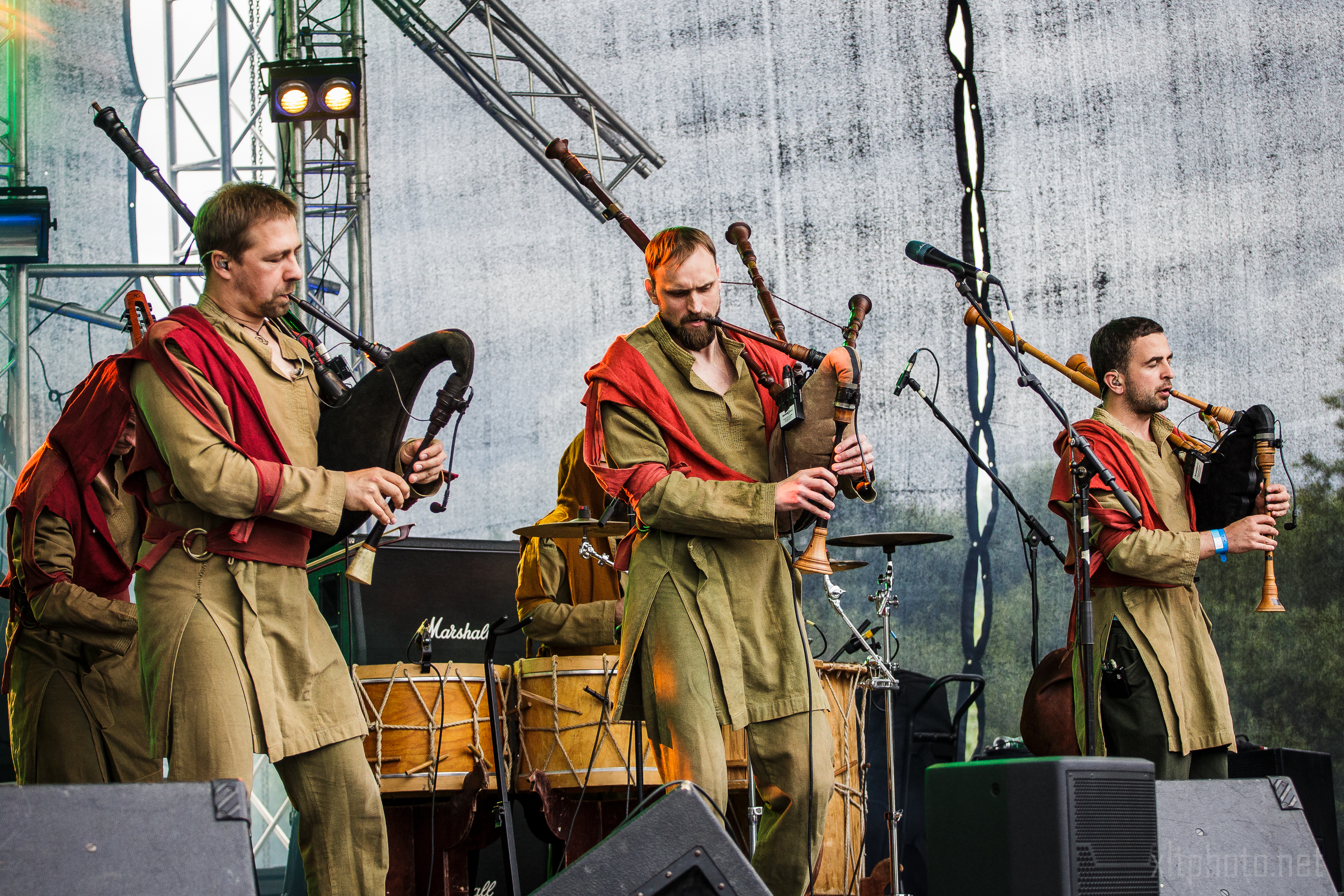 Three musicians on a stage during Kilkim Žaibu 2015 play bagpipes. They are dressed in traditional, earthy-toned outfits accented with red fabric draped around their shoulders. The background features stage equipment and a drum set, with trees faintly visible beyond the stage. Mic stands are scattered around.