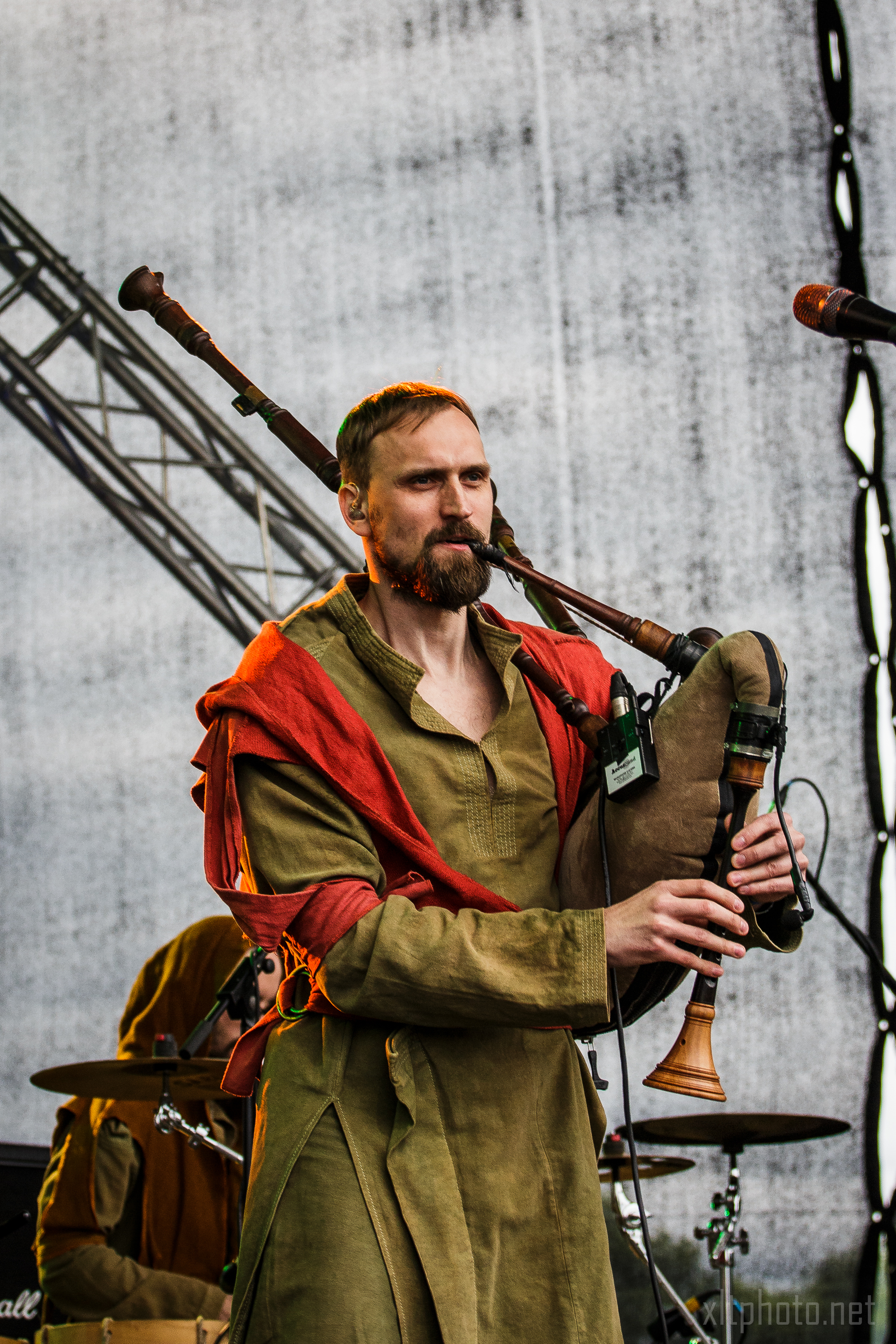 At the 2015 Kilkim Žaibu festival, a man in a green tunic and red cloak plays the bagpipes on stage with a serious expression and a trimmed beard. The background reveals stage lighting and equipment, while another person in similar attire is partially visible, playing a drum set.