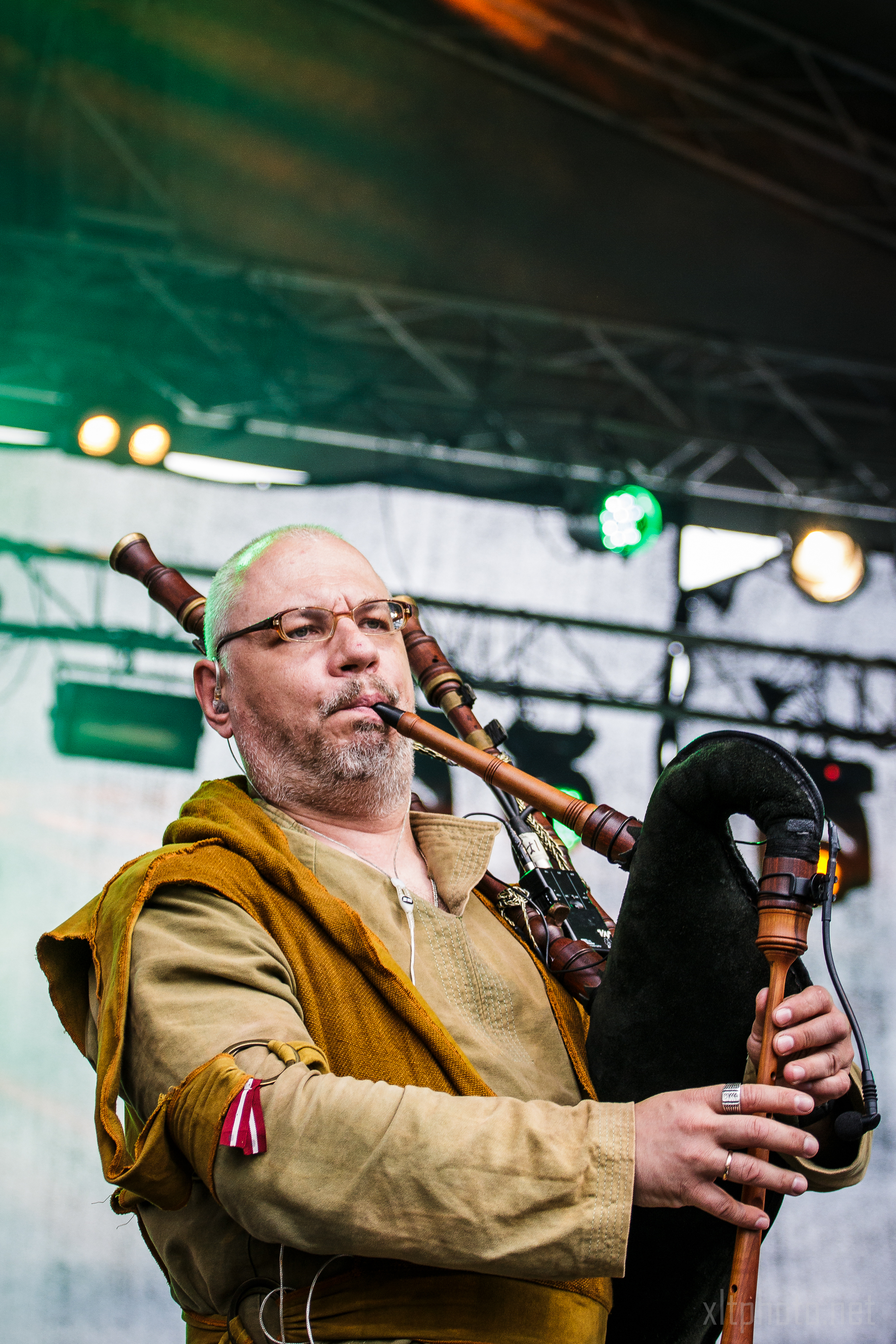 A man with glasses and a bald head is playing a bagpipe on stage during the 2015 Kilkim Žaibu festival. He is wearing a medieval-style outfit consisting of a brown tunic with green accents. The stage behind him is lit with green lights, and a truss with additional lights is visible in the background.