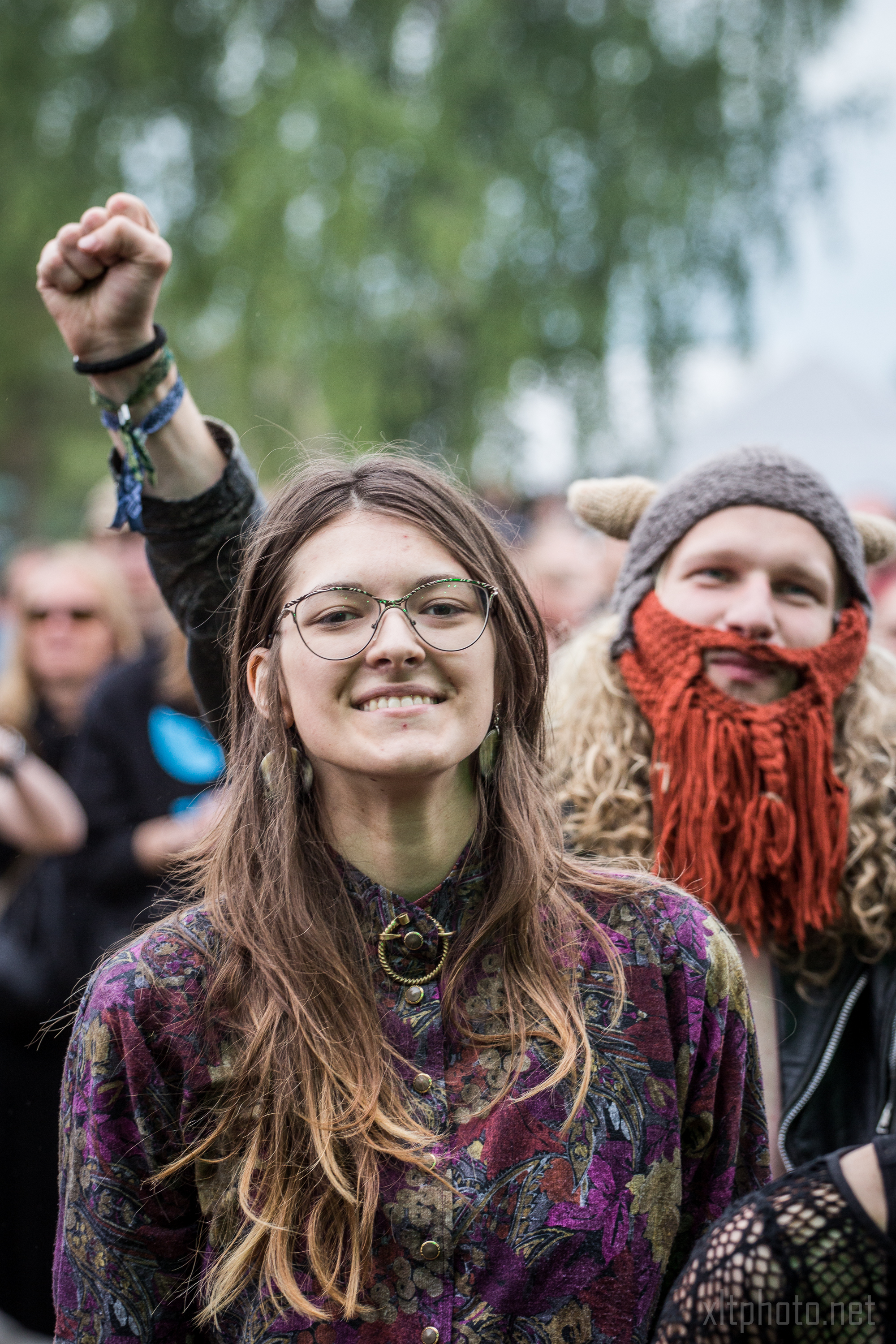 A young woman with long brown hair, glasses, and a patterned shirt stands smiling outdoors. Behind her, a person with a knitted hat resembling a Viking helmet and red beard raises a clenched fist. Blurry greenery and more people are in the background, indicating the lively atmosphere of Kilkim Žaibu 2015.