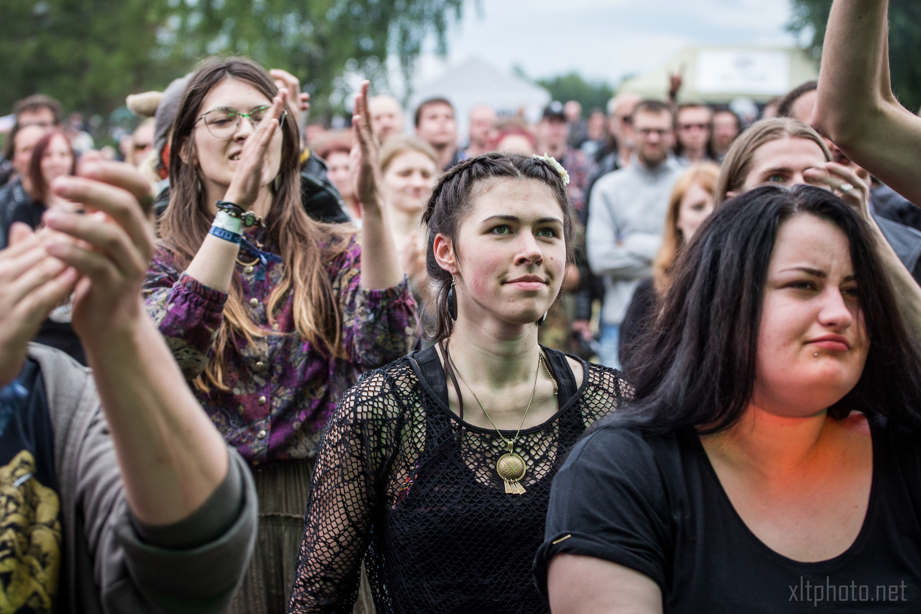 A crowd of young adults at an outdoor event, capturing the lively and engaged mood of Kilkim Žaibu 2015. One woman in the foreground has braids and wears a black netted top with a circular pendant. Another person is clapping. Trees and a white tent are visible in the background.