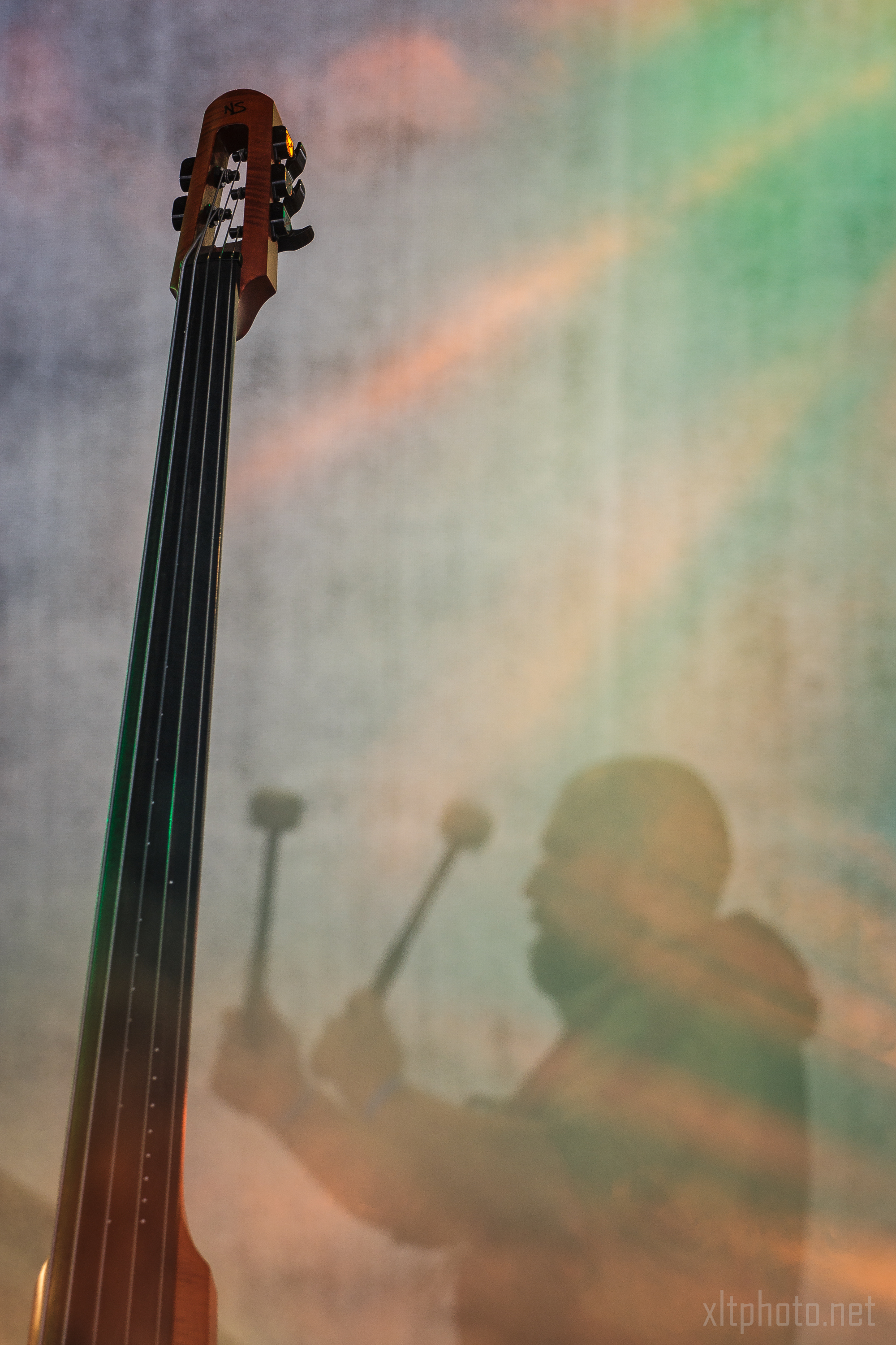 A close-up of an upright bass placed vertically in the foreground of the image. In the background, a person is playing percussion instruments with mallets, their silhouette visible against a colorful, foggy backdrop. The image has a dreamy, ethereal quality with soft lighting and hues, reminiscent of Kilkim Žaibu 2015.
