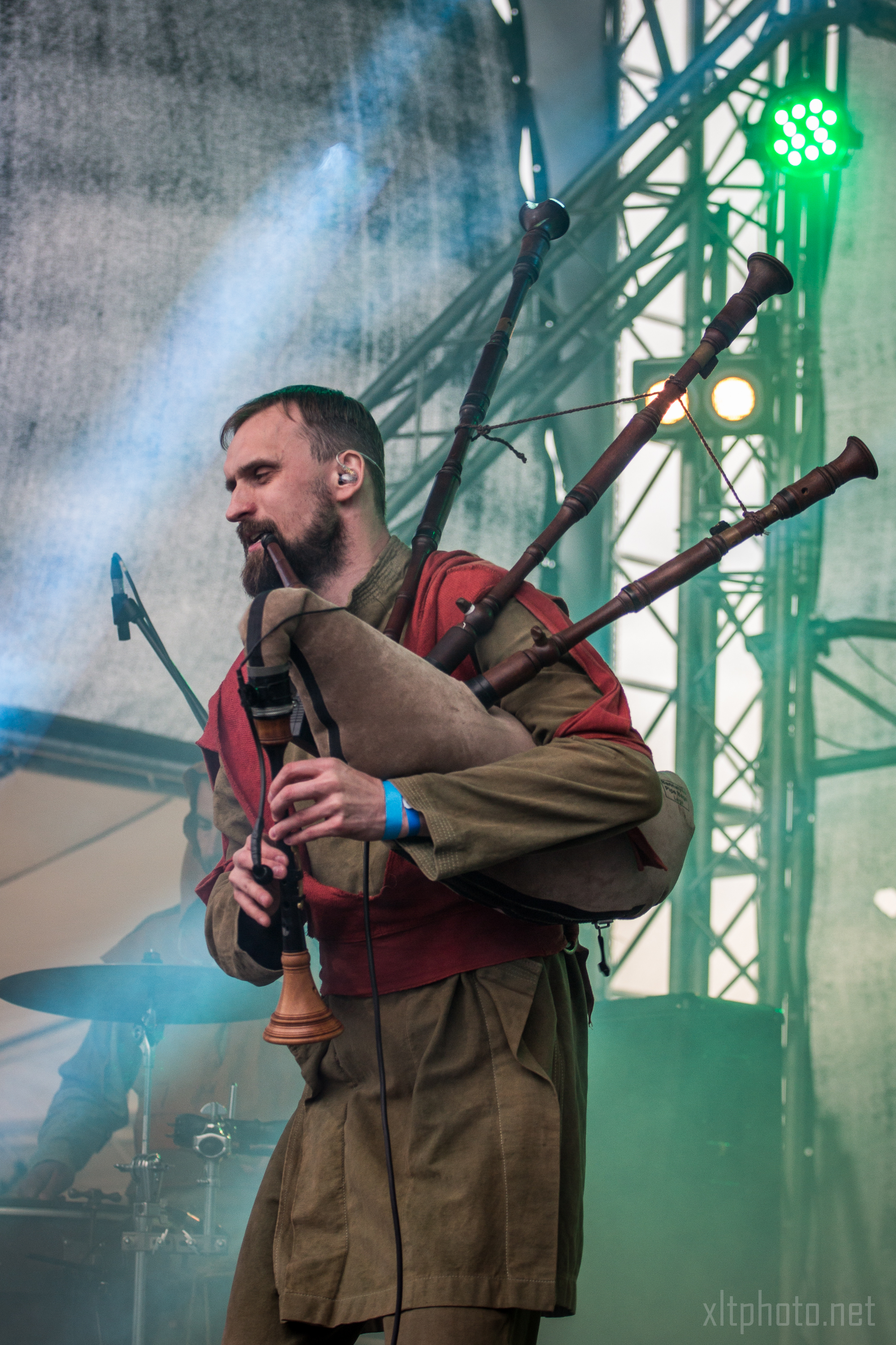 A bearded man plays the bagpipes on a stage at the 2015 Kilkim Žaibu festival, wearing a red and brown outfit. Behind him are stage lights and structural rigging. A blue wristband is visible on his right wrist. The stage is filled with smoke, and the drummer in the background is partially visible. Image credit "xltphoto.net" is in the bottom