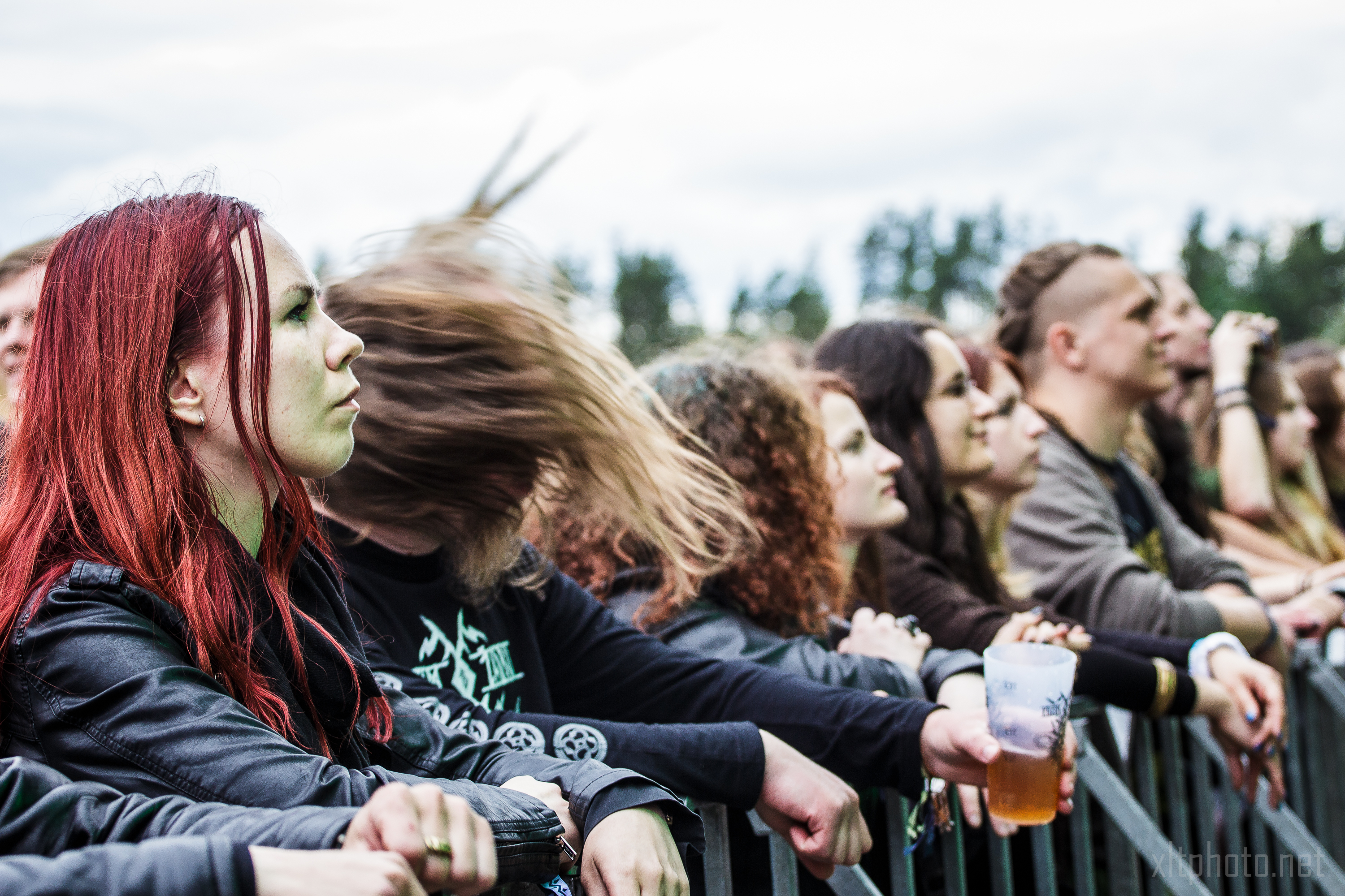 People stand behind a metal barricade at Kilkim Žaibu 2015, an outdoor concert. In the foreground, a woman with long red hair faces the stage, and a person next to her has their hair flying wildly. Many attendees focus on the performance, with one individual holding a beer cup. Trees blur in the background.