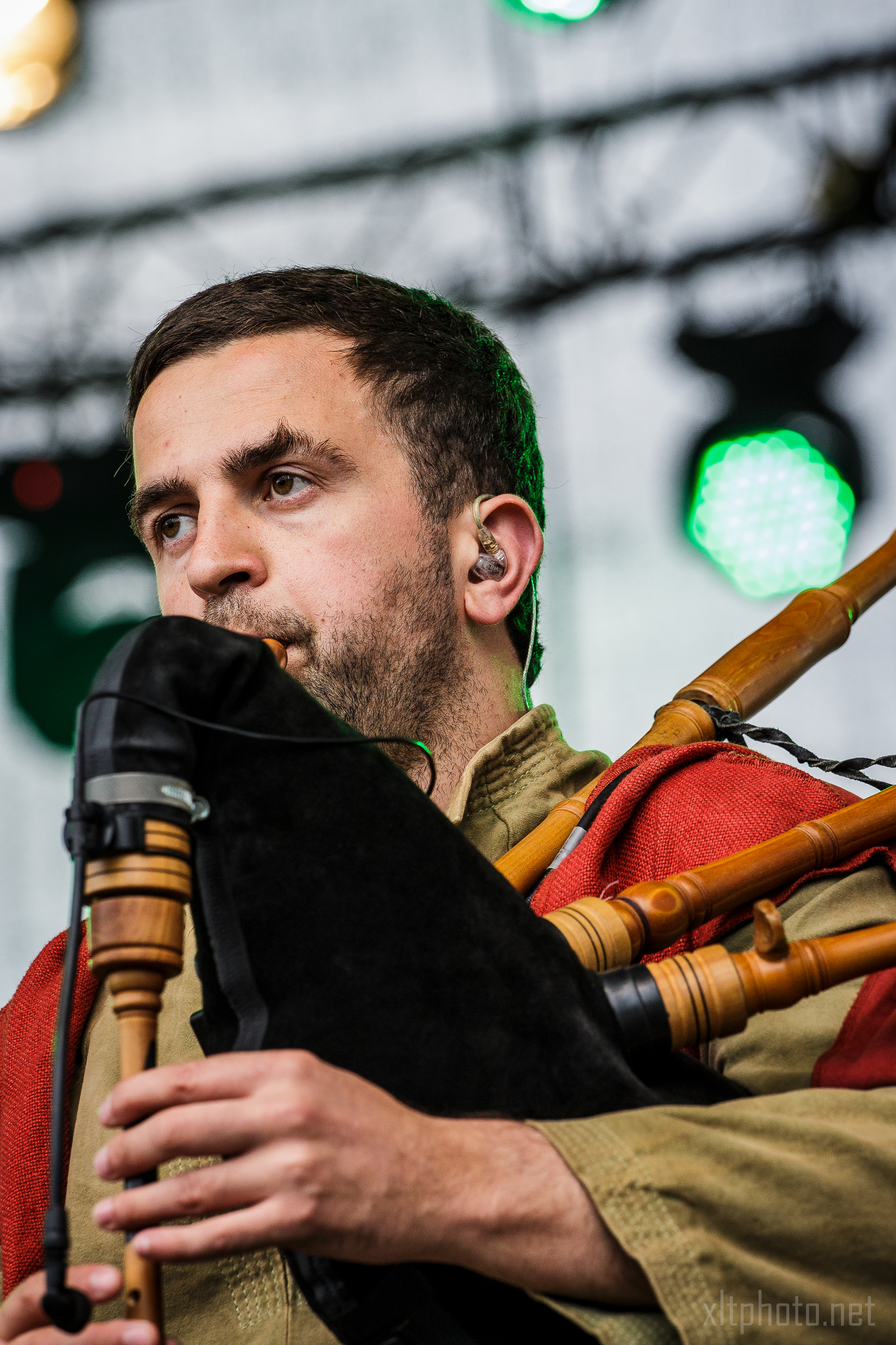 A man with short hair and a trimmed beard plays a bagpipe on stage at Kilkim Žaibu 2015. He wears in-ear monitors and is dressed in a green outfit with an orange-red shawl over his shoulder, performing amidst blurred lights and equipment in the background.