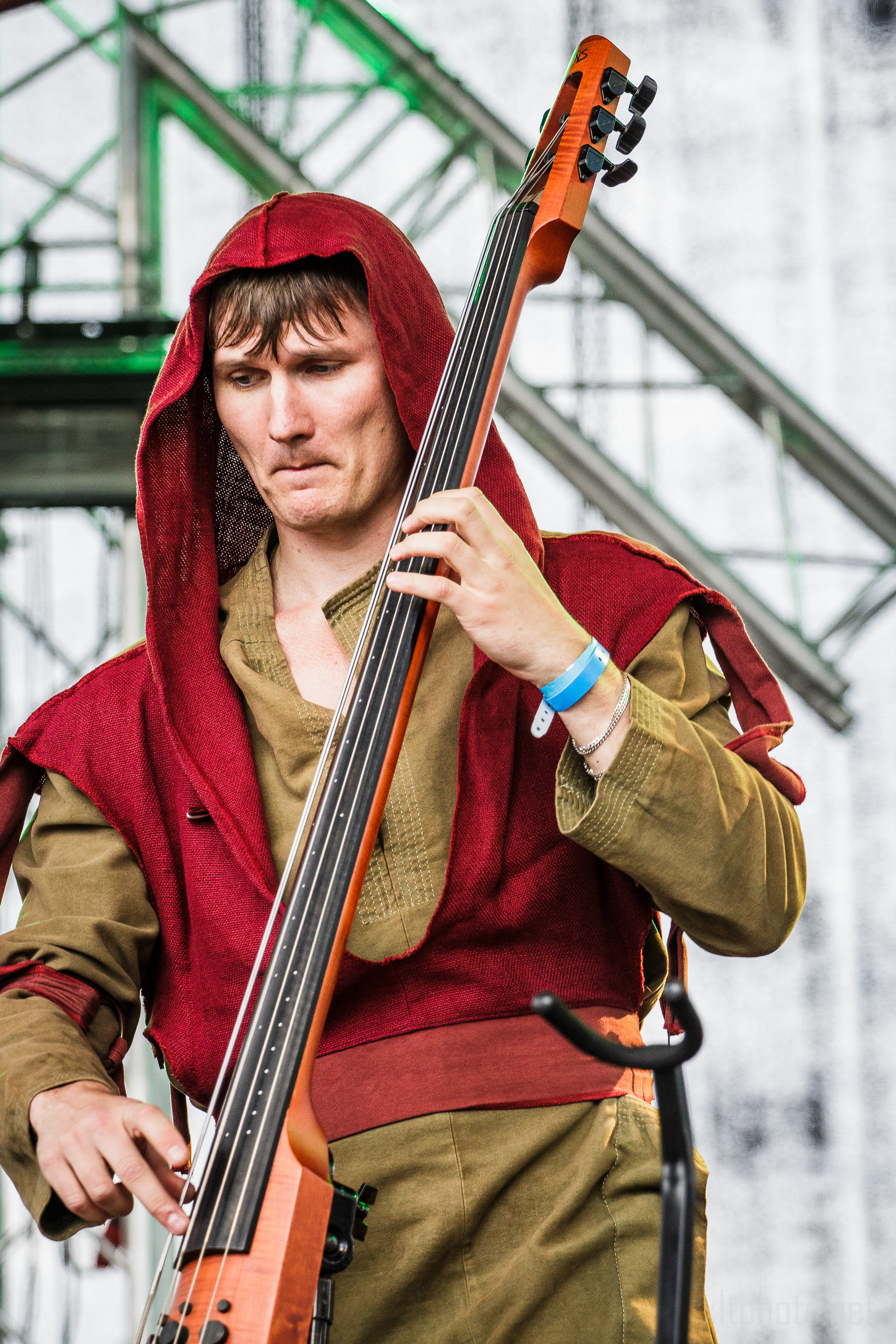 A man is playing an upright electric bass on stage at the 2015 Kilkim Žaibu festival. He is wearing a medieval-style outfit with a red hooded cloak over a green tunic and pants. The background features stage lighting and metal scaffolding. He has a wristband on his right wrist and appears focused on his performance.
