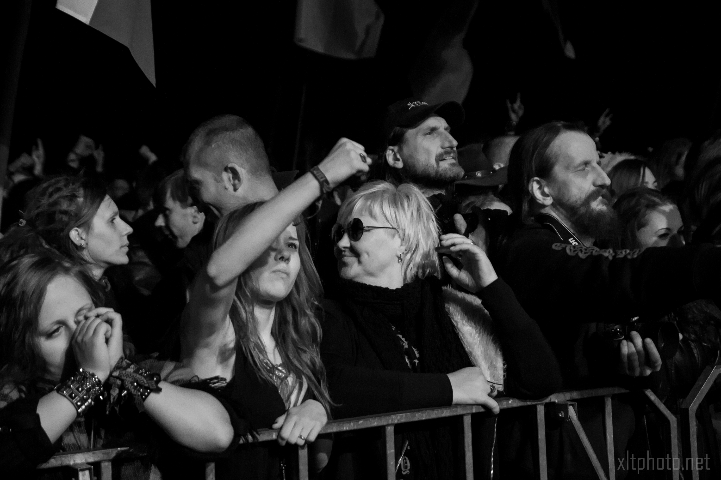 A black and white image captures the energy of the Kilkim Žaibu festival in 2015. The outdoor concert crowd is tightly packed at night, with some hands raised. The central figure, wearing sunglasses, smiles and looks right. Another person with a beard and long hair stands to the right, as flags wave in the background.