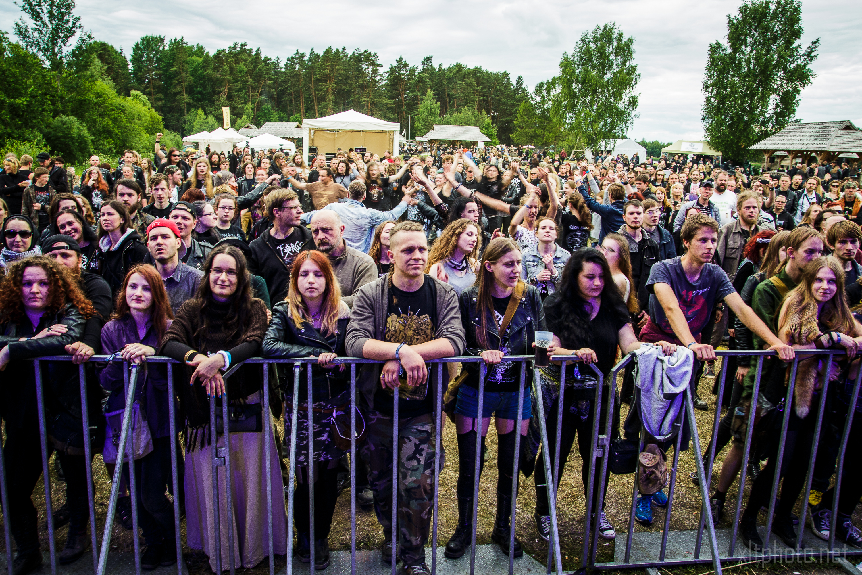 A large crowd gathers outdoors at the Kilkim Žaibu event, standing behind metal barricades. The attendees, wearing various styles of clothing including band shirts and jackets, are actively watching a 2015 performance. Trees and tents are visible in the background under a cloudy sky.