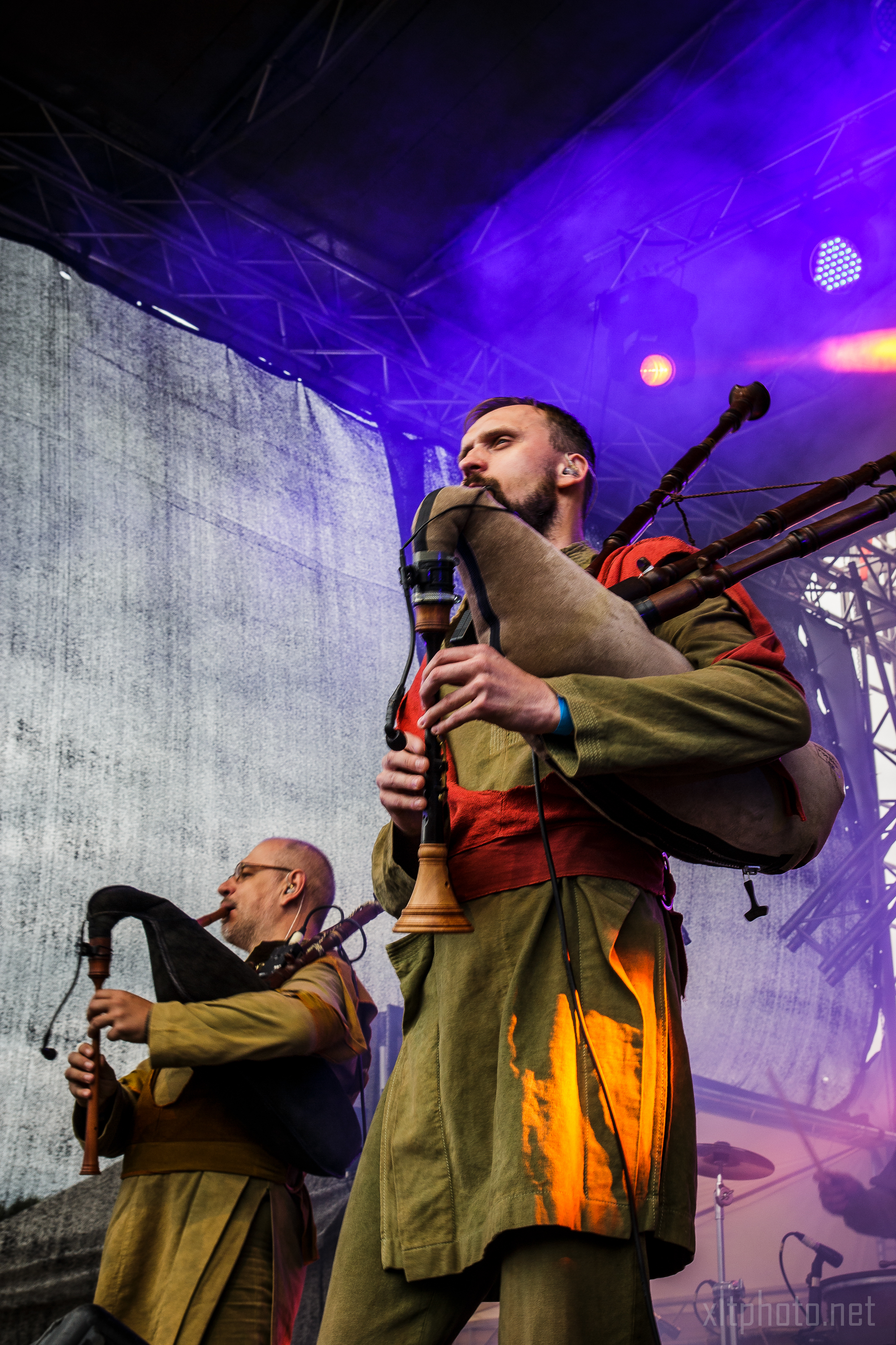 Two musicians perform on stage with bagpipes at what seems to be a festival. The man in the foreground, wearing a green robe and red sash, plays intently, while the man in the background also focuses on his playing. The stage is bathed in moody purple and blue lighting, with a large backdrop screen behind them.
