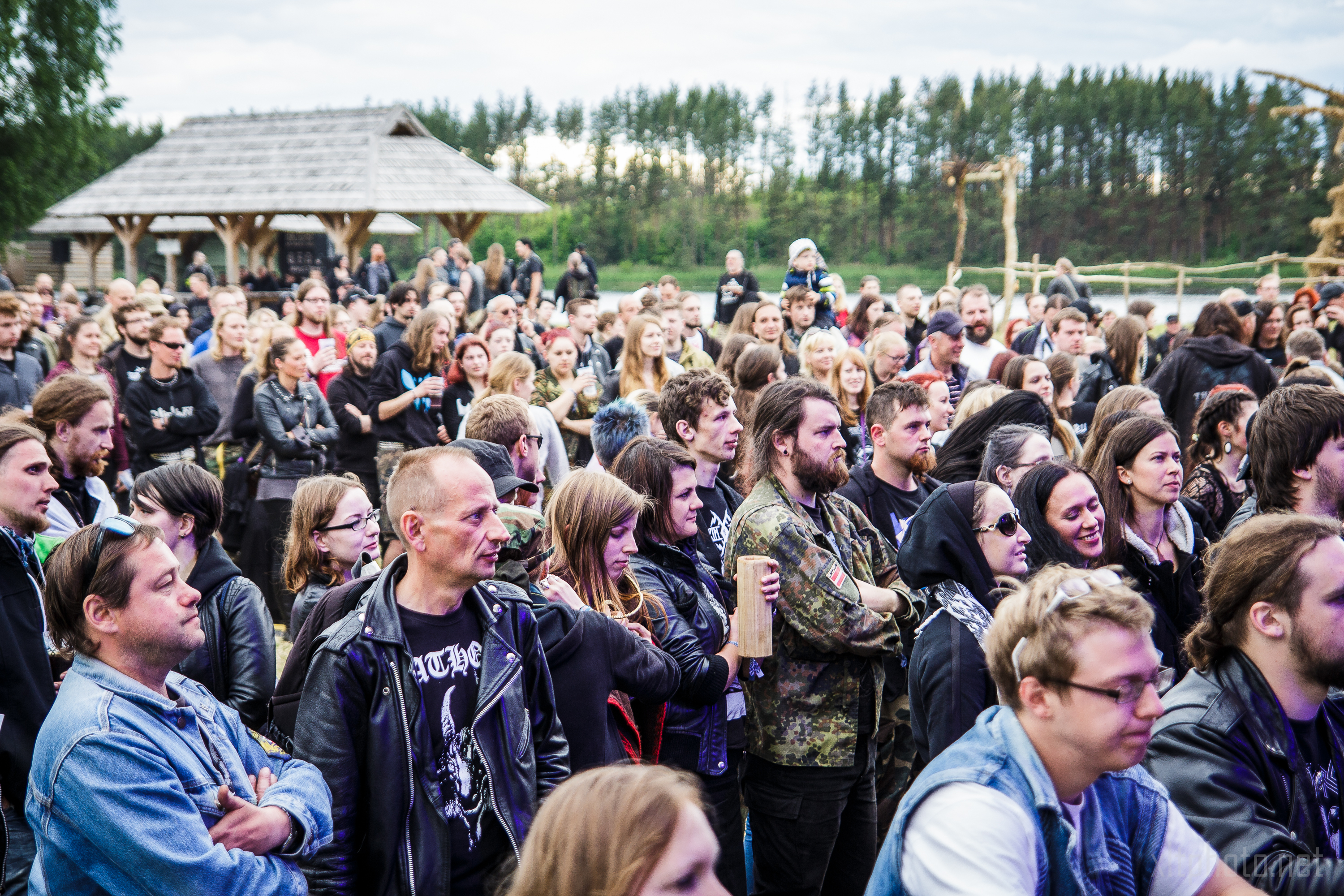 A large crowd of people, predominantly dressed in dark clothing and leather jackets, are gathered outdoors for Kilkim Žaibu 2015. The background features a wooded area with green trees and a rustic wooden shelter. Attendees appear to be enjoying the event under a cloudy sky.