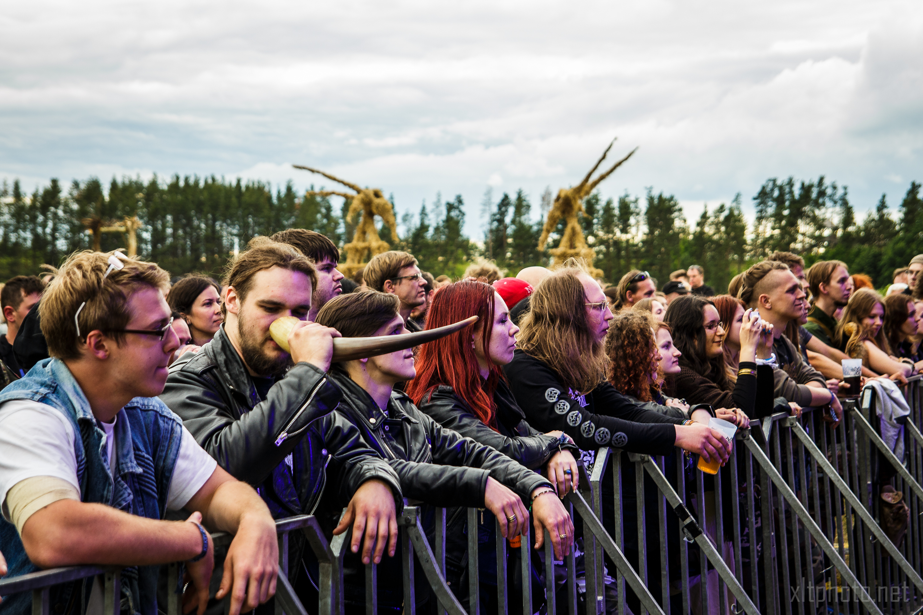 A crowd at the 2015 Kilkim Žaibu festival is gathered behind a metal barrier. A man in the foreground holds and blows into a horn. Many attendees have long hair and wear dark clothing. Trees and large wooden structures are visible in the background under a cloudy sky.