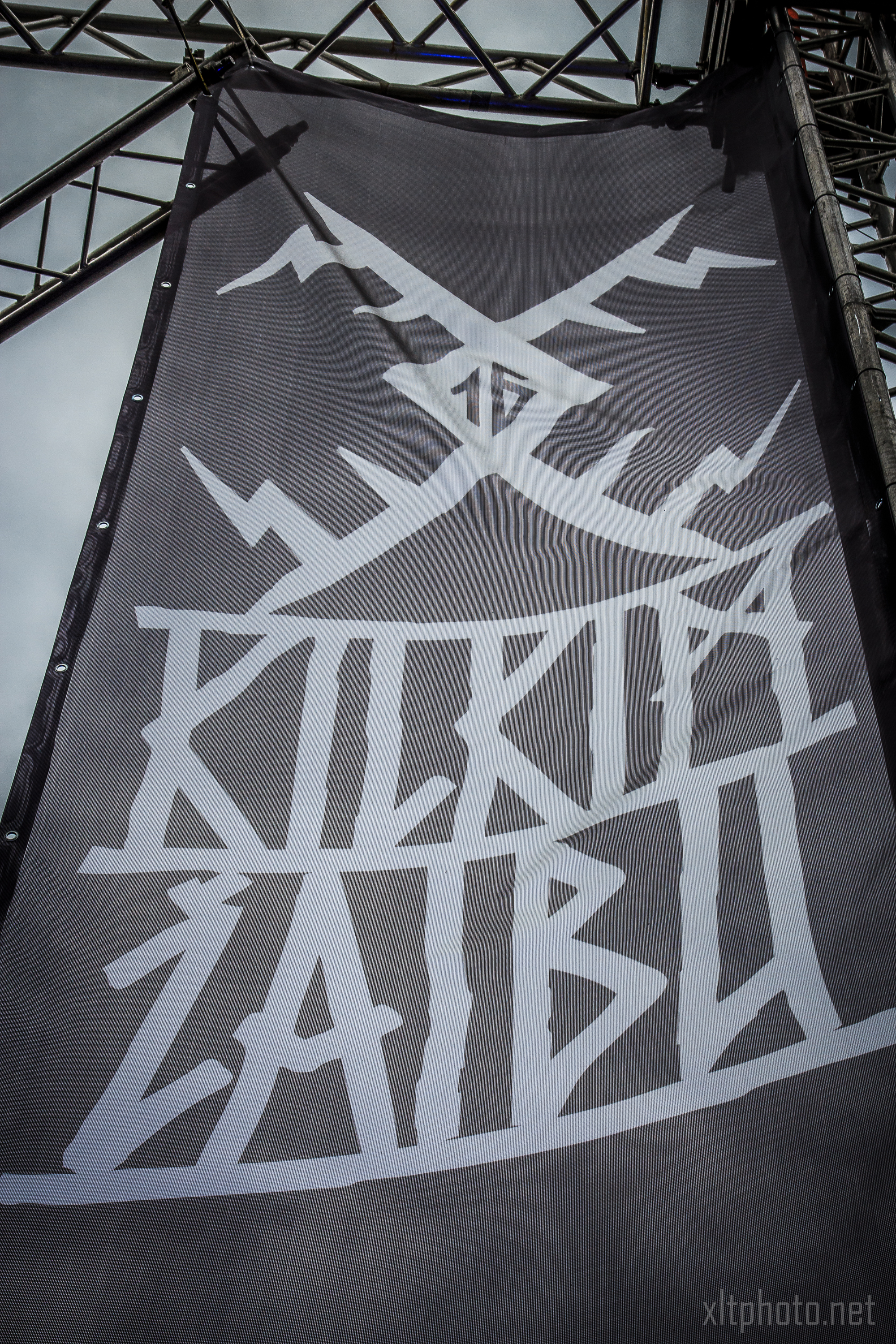 A large black banner with abstract, angular white designs and text hangs between metal scaffolding against a cloudy sky. The stylized text may spell "KILKIM ŽAIBU 2015." The lower right corner of the image has the watermark "xltphoto.net.