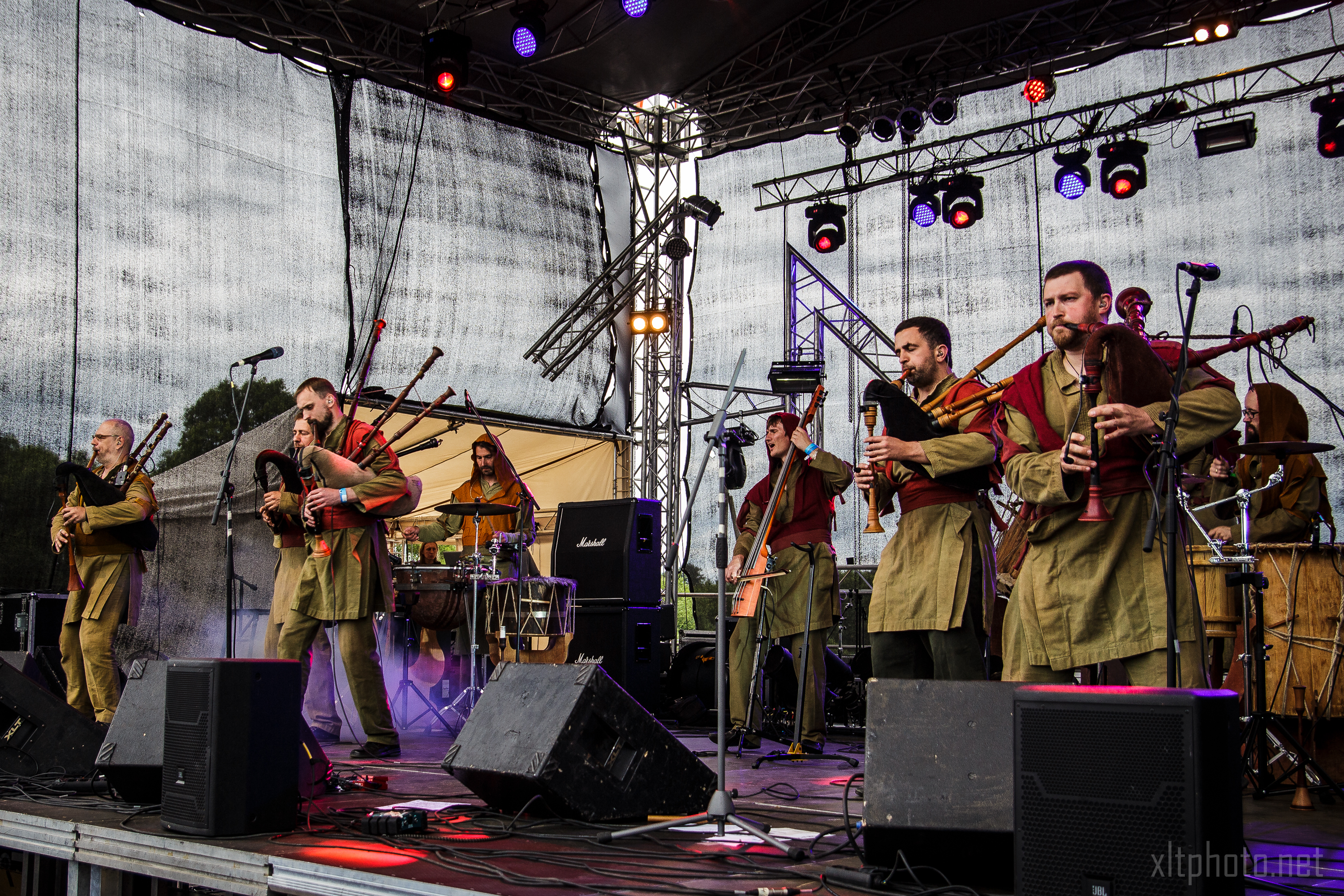 At the 2015 Kilkim Žaibu festival, a band of musicians dressed in earth-toned medieval-style clothing performs on an outdoor stage. Some play bagpipes while others beat drums. Colorful lights illuminate the scene, and various speakers and equipment are visible amidst a large netted backdrop surrounding the stage area.
