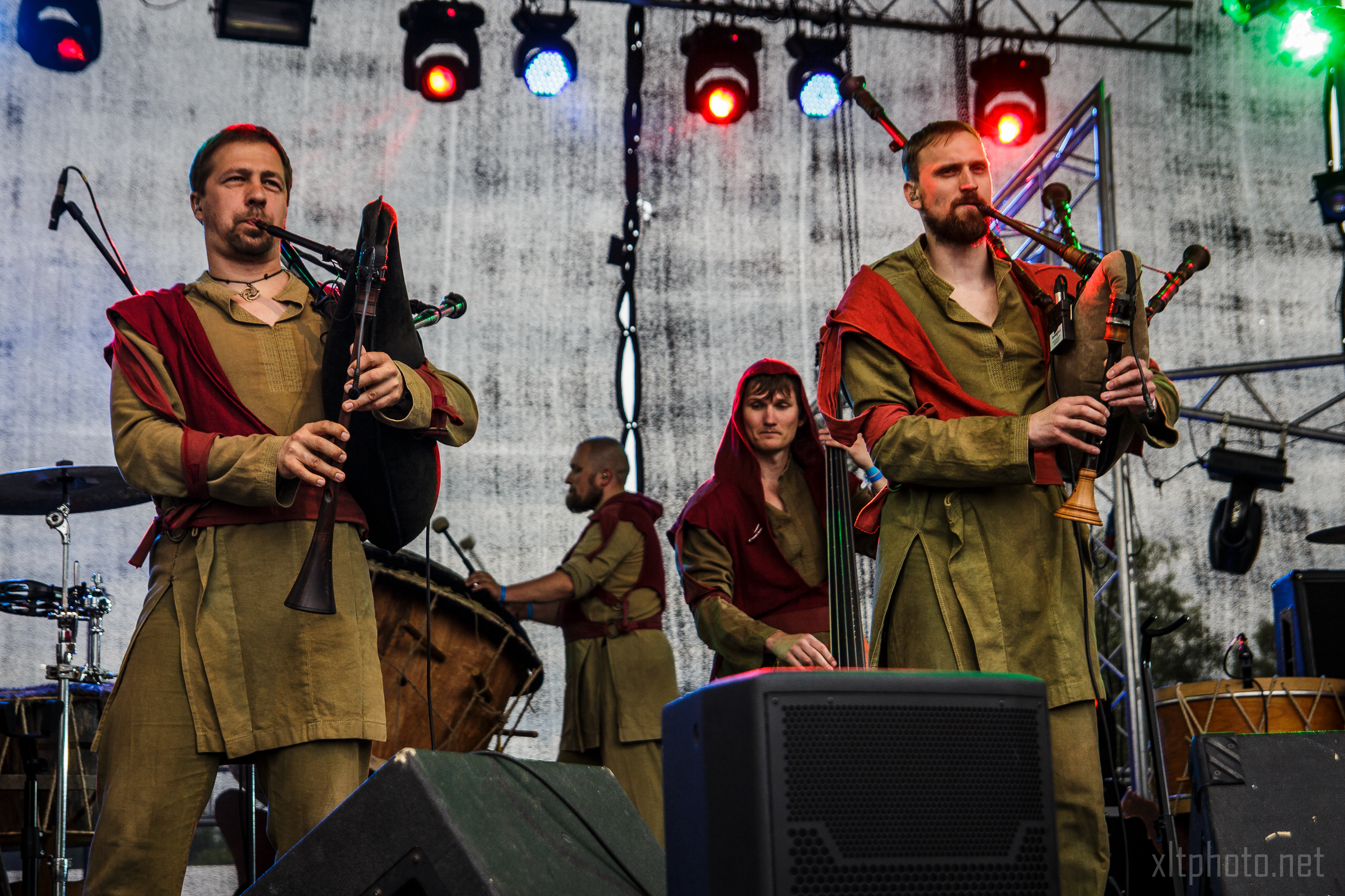 A group of musicians dressed in medieval-style tunics with red scarves play bagpipes and drums on a stage. The backdrop, reminiscent of the vibrant atmosphere at Kilkim Žaibu 2015, is lit with colorful lights, and musical equipment is visible in the foreground. A woman in the center appears to adjust an instrument.