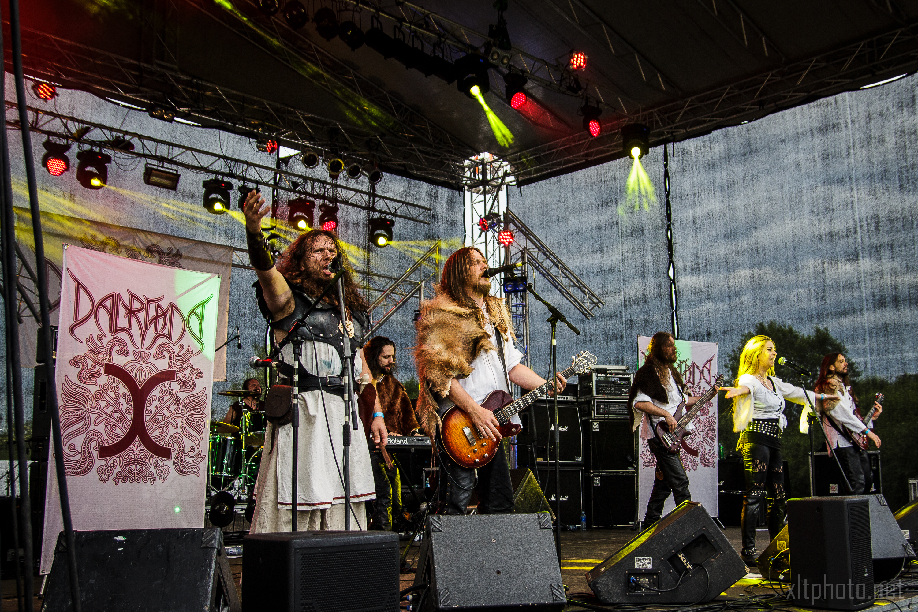 A band performs on stage under colorful lights at the 2015 Kilkim Žaibu outdoor event. The frontman, dressed in medieval-inspired clothing, raises his hand while another musician plays a guitar. Other band members are visible, and the backdrop features banners with artistic designs. Equipment and speakers are visible in the background.
