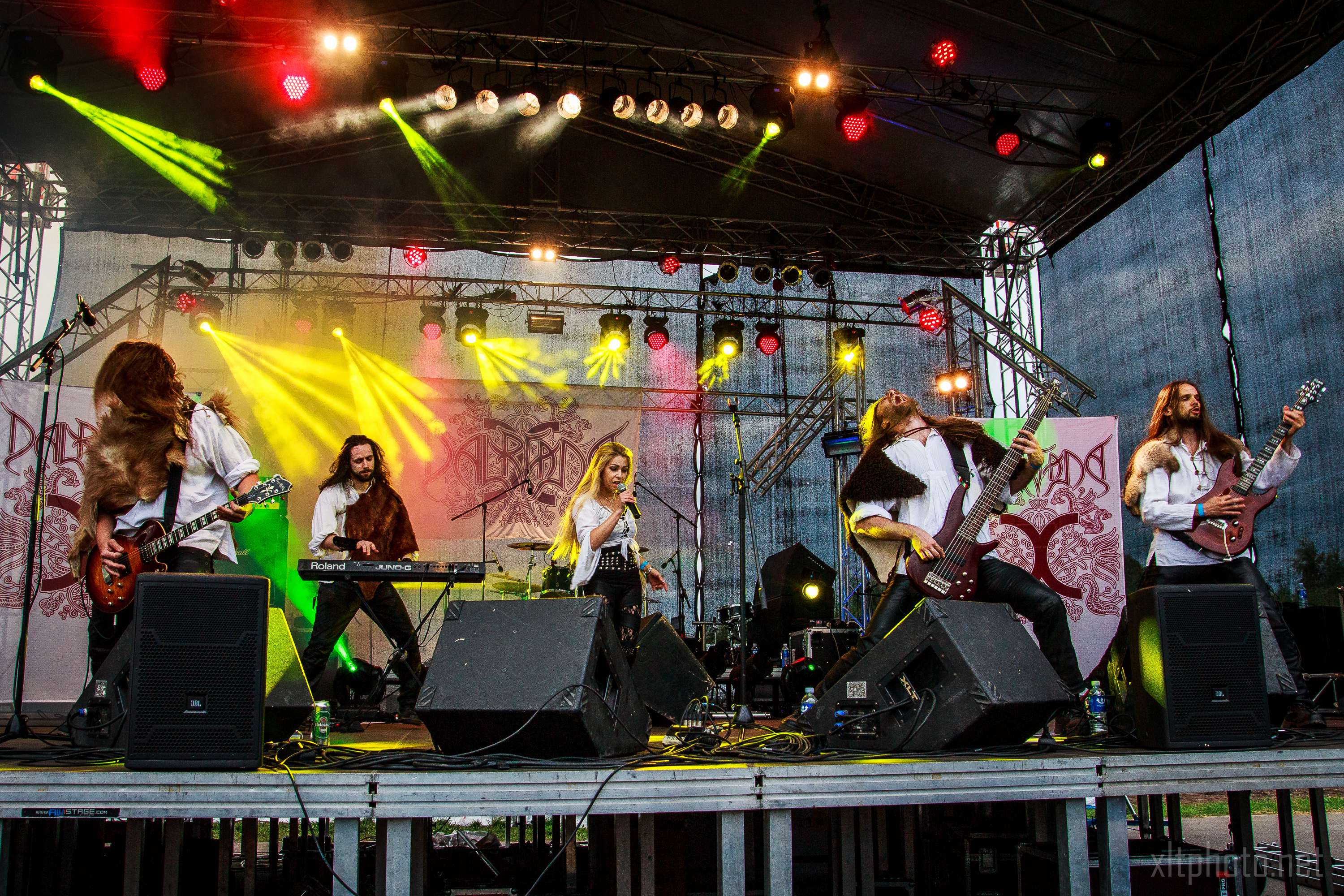 A live band performs on stage with dynamic lighting effects reminiscent of Kilkim Žaibu 2015. The lineup includes a keyboardist, a female vocalist, and three male guitarists. The stage is adorned with red and yellow lights, and a backdrop featuring the band's logo. The musicians are energetically engaged in their performance.