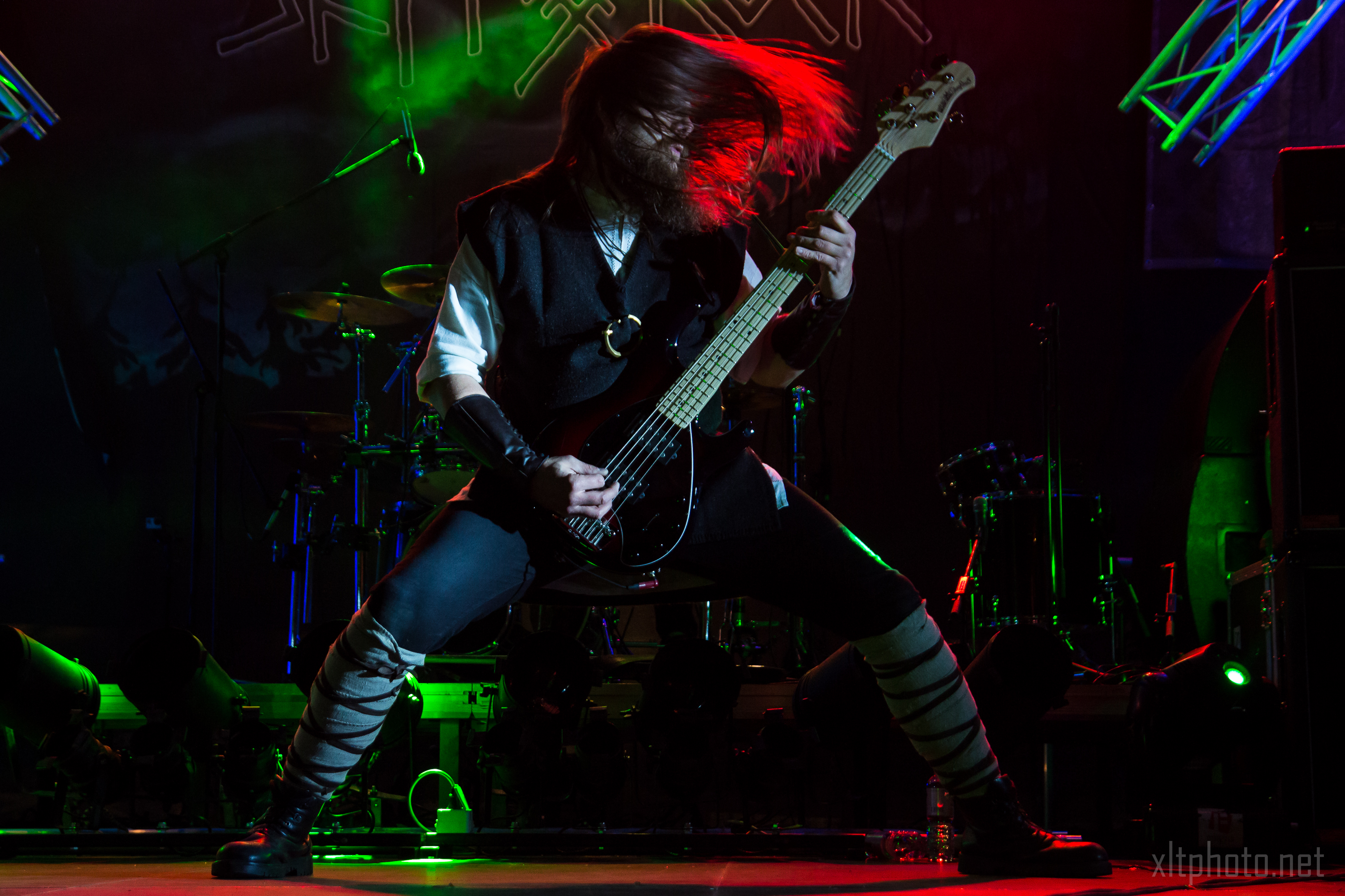 A musician with long hair thrashes their head while playing a black electric guitar on stage at Kilkim Žaibu 2015. They are wearing a dark outfit with light accents on the legs and arms. The background is dimly lit with green and purple lights, showcasing musical instruments and equipment.