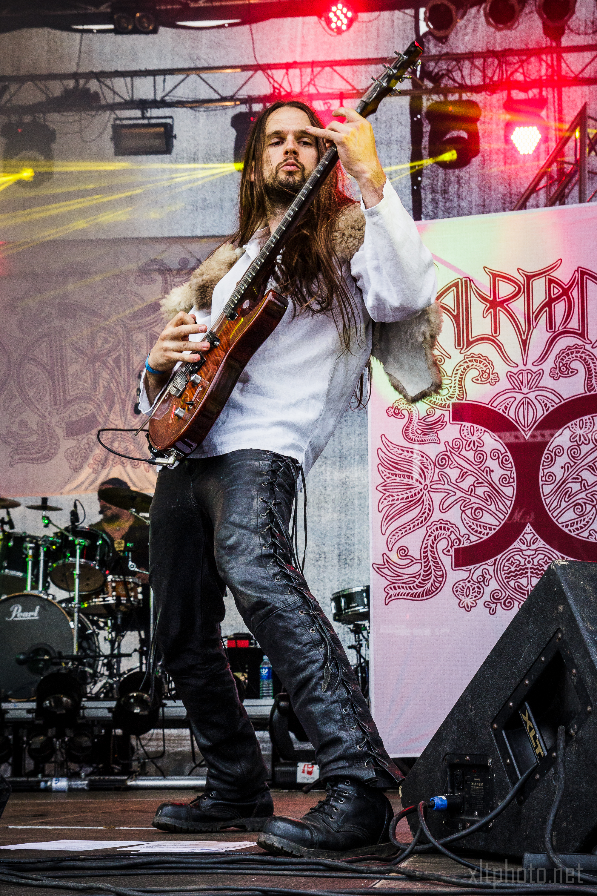 A musician with long hair plays an electric guitar onstage at the 2015 Kilkim Žaibu festival. He wears a white shirt and black leather pants with side lace detailing. In the background, a drummer plays a drum kit. The stage is decorated with ornate artwork, and colorful lights illuminate the scene.