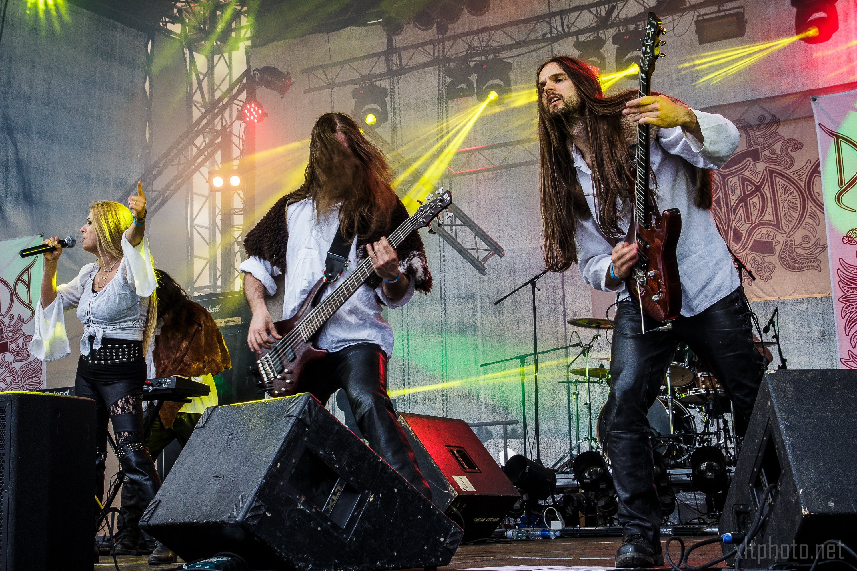 A rock band performs energetically on stage at the 2015 Kilkim Žaibu festival. The female singer points towards the crowd while two male guitarists, both with long hair, play intensely. Bright stage lights and colored beams create a dynamic atmosphere. A drummer is visible in the background. Band's name is partially visible on the backdrop.