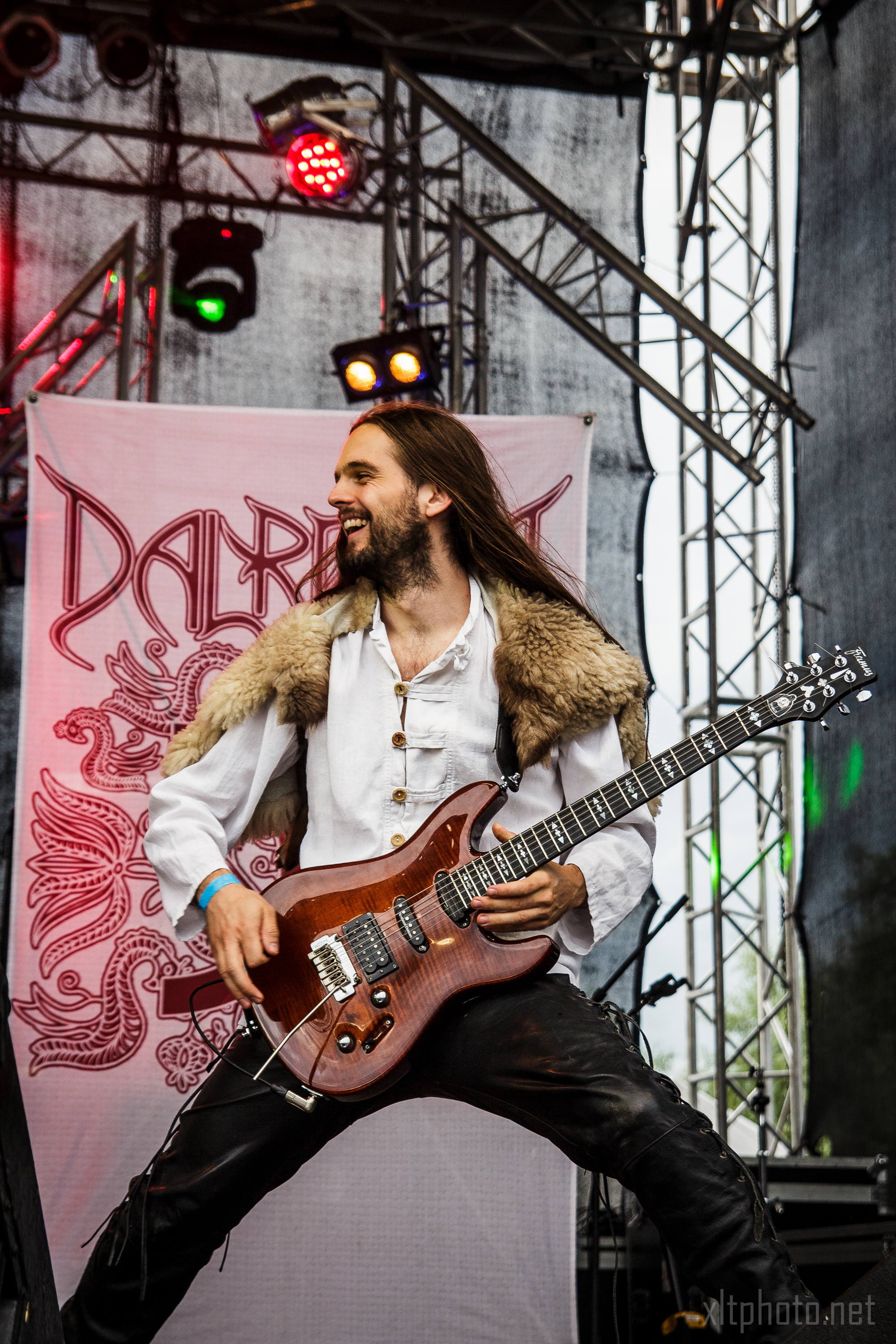 A musician with long hair, wearing a white shirt and fur vest, plays an electric guitar on stage at Kilkim Žaibu festival. Colorful stage lights illuminate him, and a banner with intricate red designs is in the background. He appears energetic and engaged with the performance, smiling broadly.