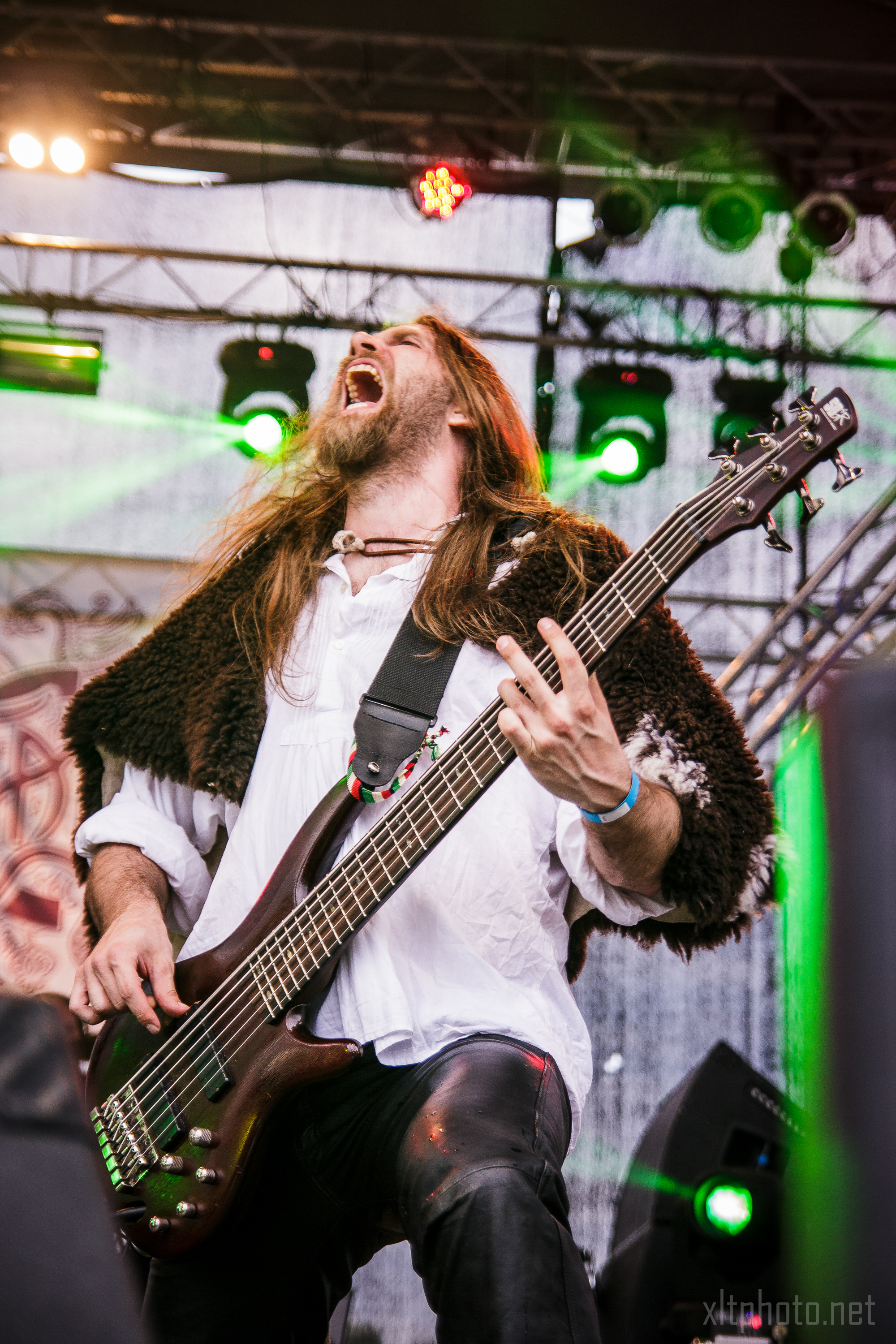 A musician with long hair and a beard passionately plays an electric bass guitar on stage at the 2015 Kilkim Žaibu festival. He is wearing a white shirt and black pants with a fur garment over his shoulders. Colorful stage lights and scaffolding are in the background, and the musician's mouth is wide open as if singing or shouting.