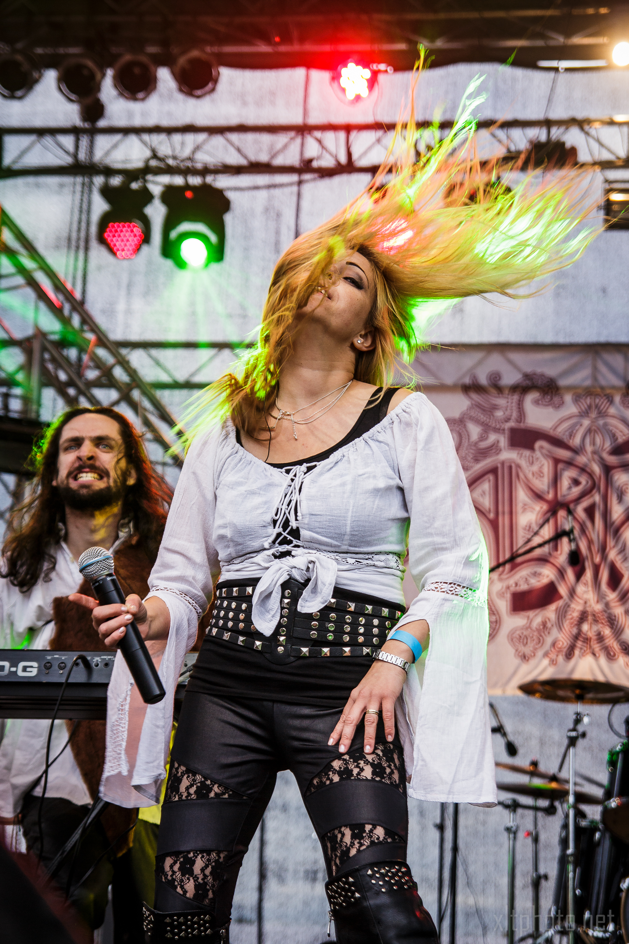 A woman on stage at the 2015 Kilkim Žaibu festival, with long hair flying mid-air, is energetically performing. She is wearing a white blouse, black pants with lace cutouts, and a studded belt. A man in the background is playing a keyboard. The stage is lit with red and green lights against an intricate backdrop.