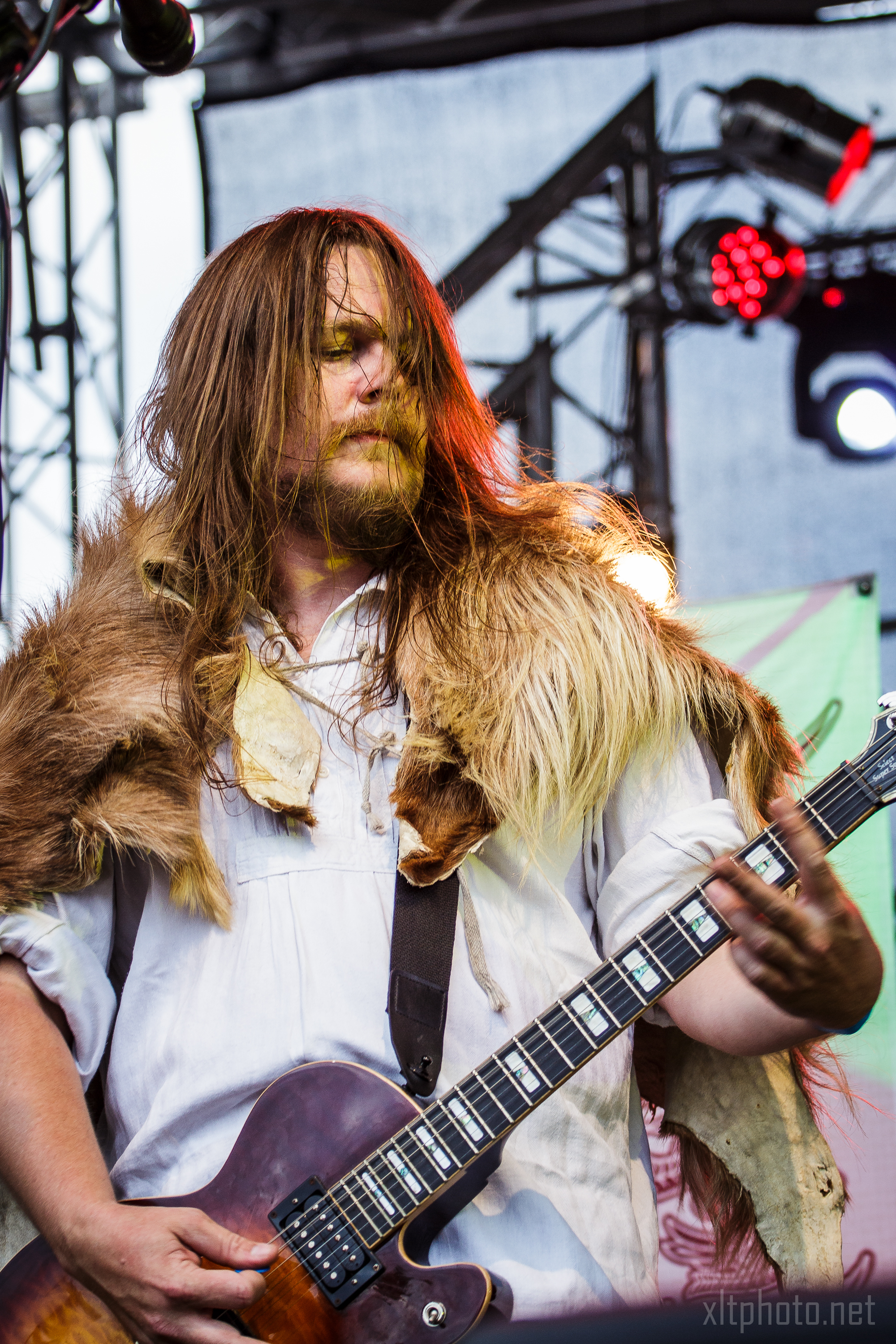 A musician with long hair performs on stage at Kilkim Žaibu 2015, playing an electric guitar. He is wearing a fur cloak over a white shirt. The stage has metal scaffolding and lights in the background. The atmosphere is vibrant, with a focus on his intense expression and dynamic posture.
Credit: xltphoto.net.