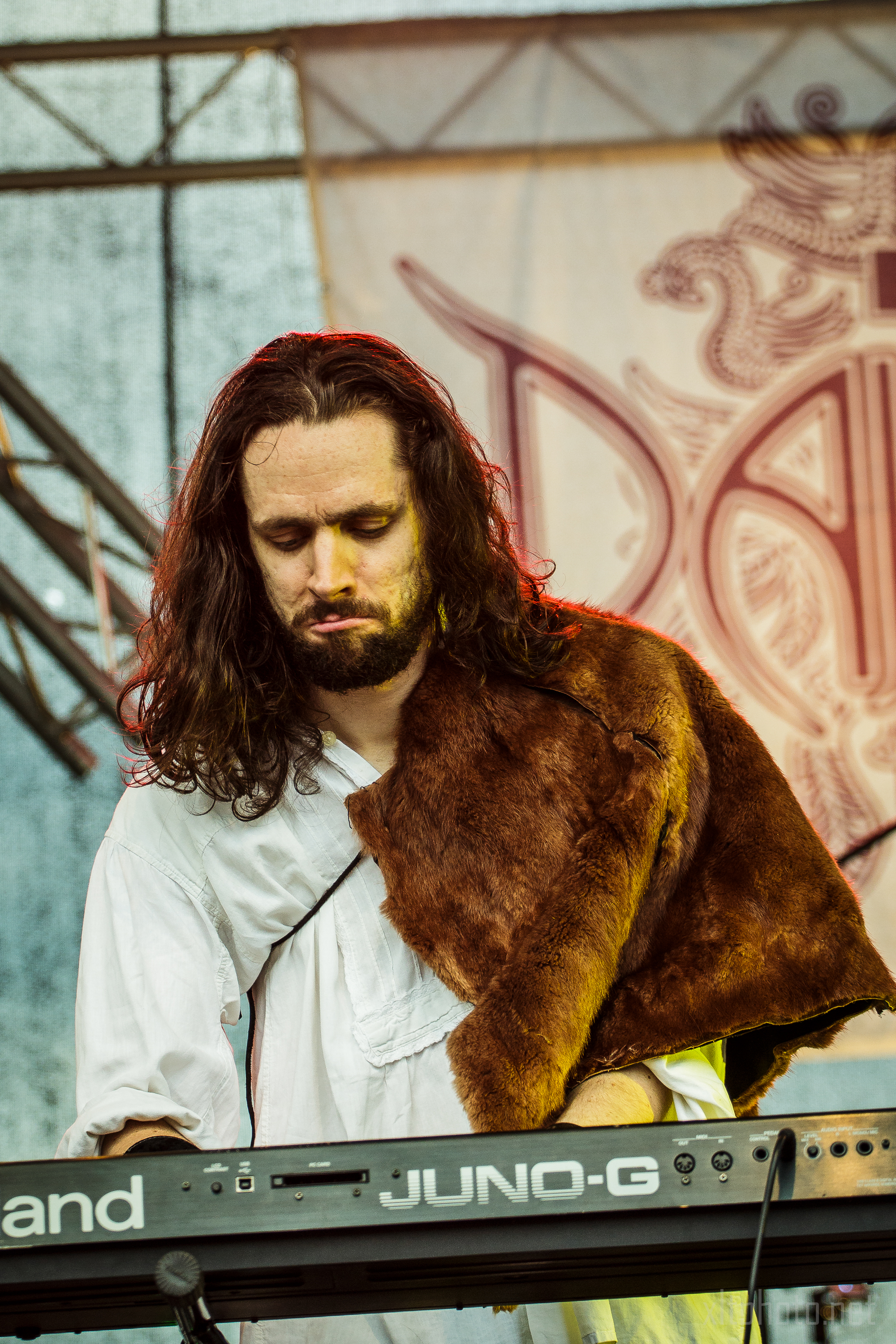 A musician with long, dark hair and a beard plays a Roland Juno-G synthesizer on stage at Kilkim Žaibu 2015. He wears a white shirt and a brown fur stole over one shoulder. The background features part of a large emblem or logo with intricate designs and the partial letters "DA".