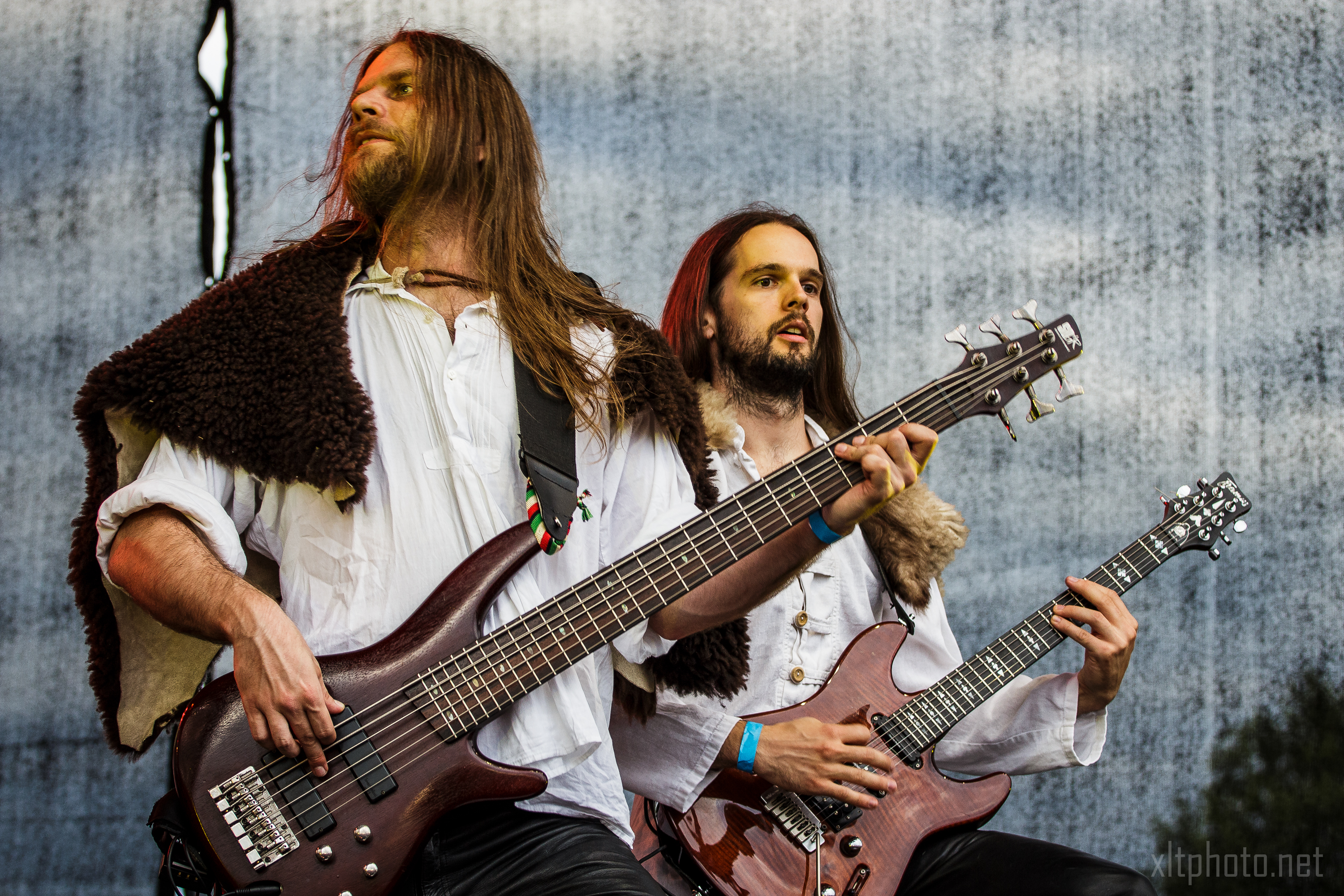 At the 2015 Kilkim Žaibu festival, two musicians with long hair, dressed in medieval-style white shirts and fur cloaks, are playing electric guitars on an outdoor stage. The foreground musician plays a bass guitar, while the background musician plays a rhythm guitar, both deeply focused on their performance.