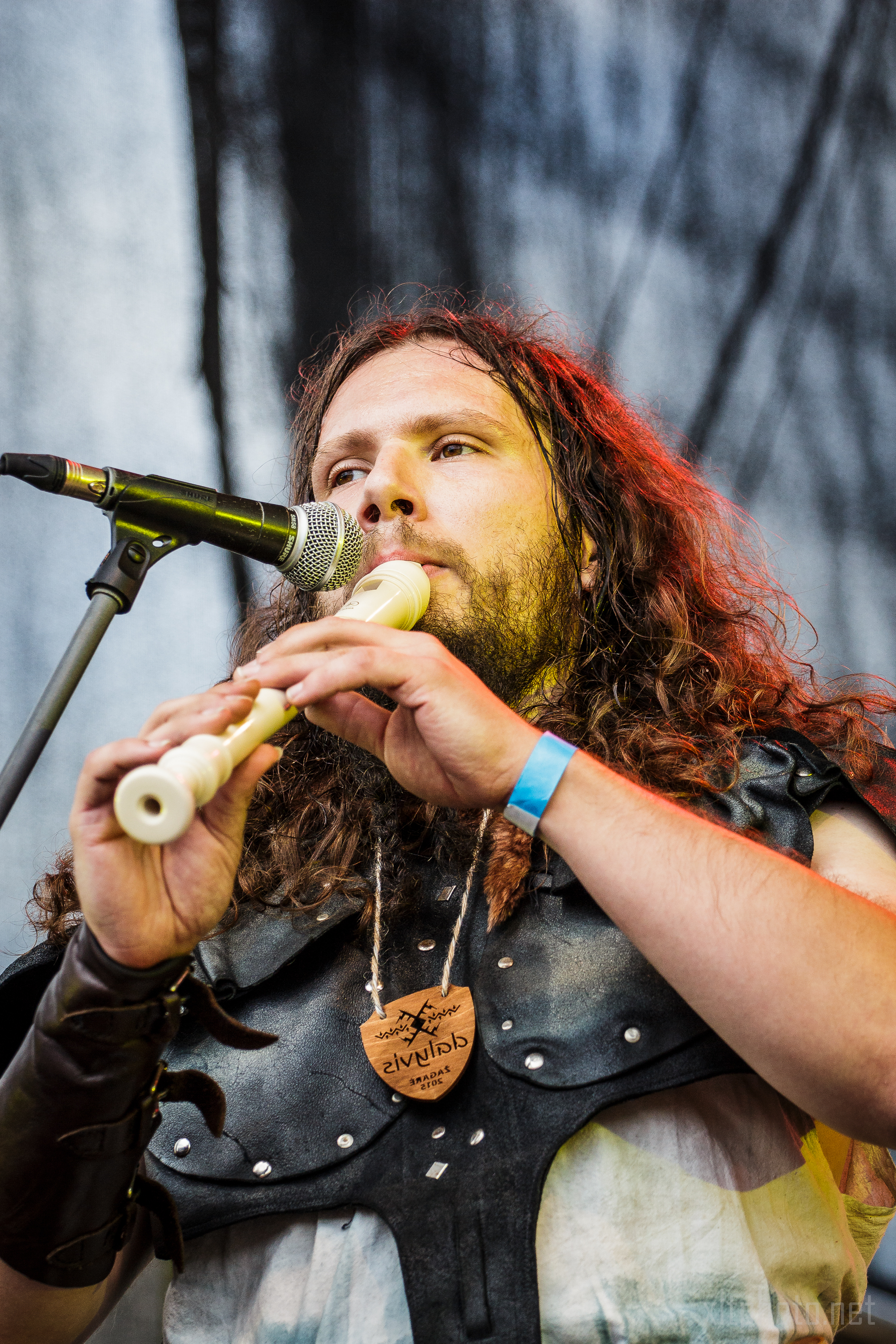At the 2015 Kilkim Žaibu festival, a man with long curly hair plays a recorder into a microphone on stage. He is wearing a medieval-inspired outfit with a leather armband and a blue wristband on his right arm. The background features a blurred, dark stage curtain. He has a focused expression and is facing slightly to the right.