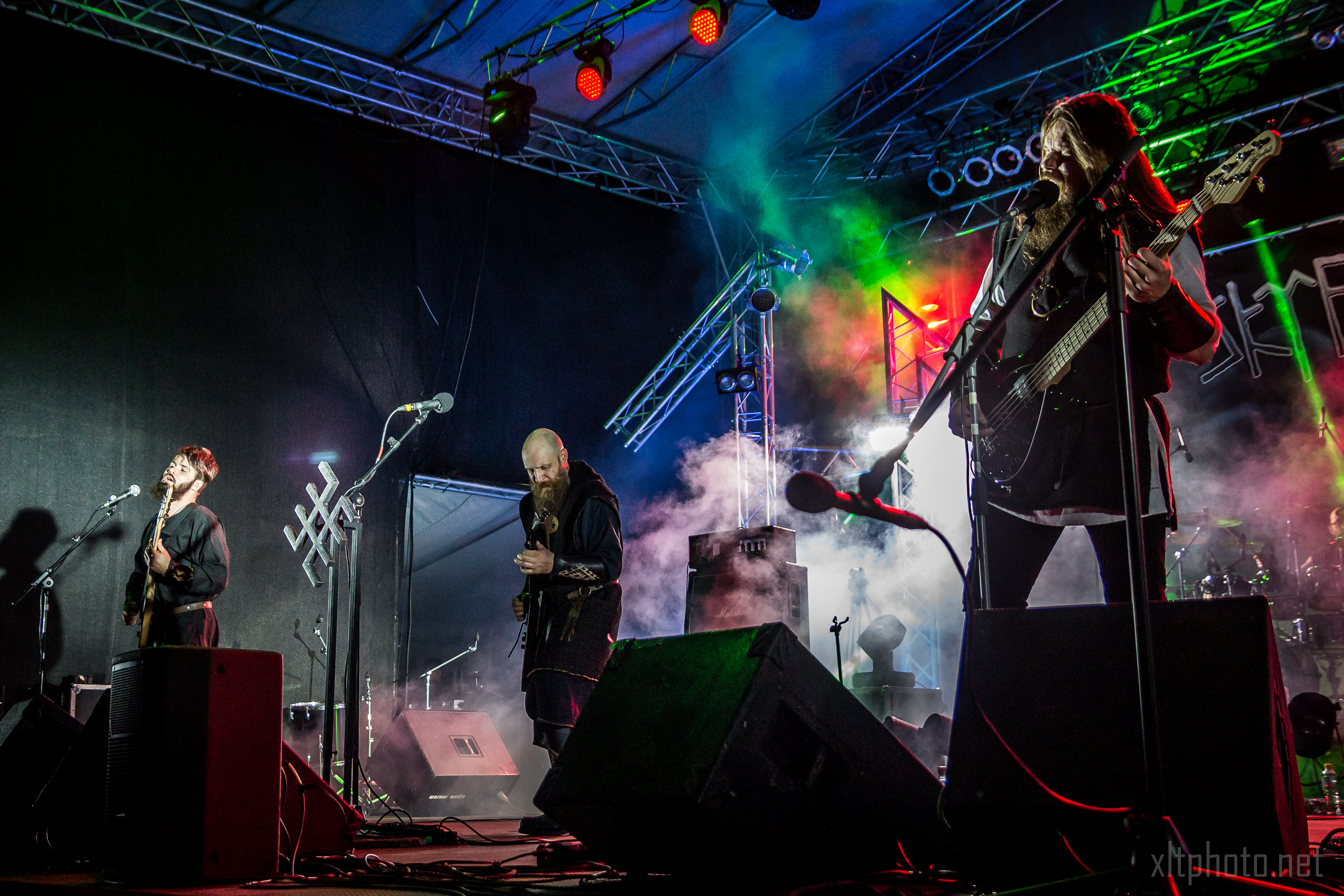 A live music performance at the Kilkim Žaibu 2015 festival features three musicians on stage. They are illuminated by vibrant colored lights with fog enhancing the visual atmosphere. The musicians are playing guitars and appear deeply engaged in their performance. The stage has a metal frame and various sound equipment.
