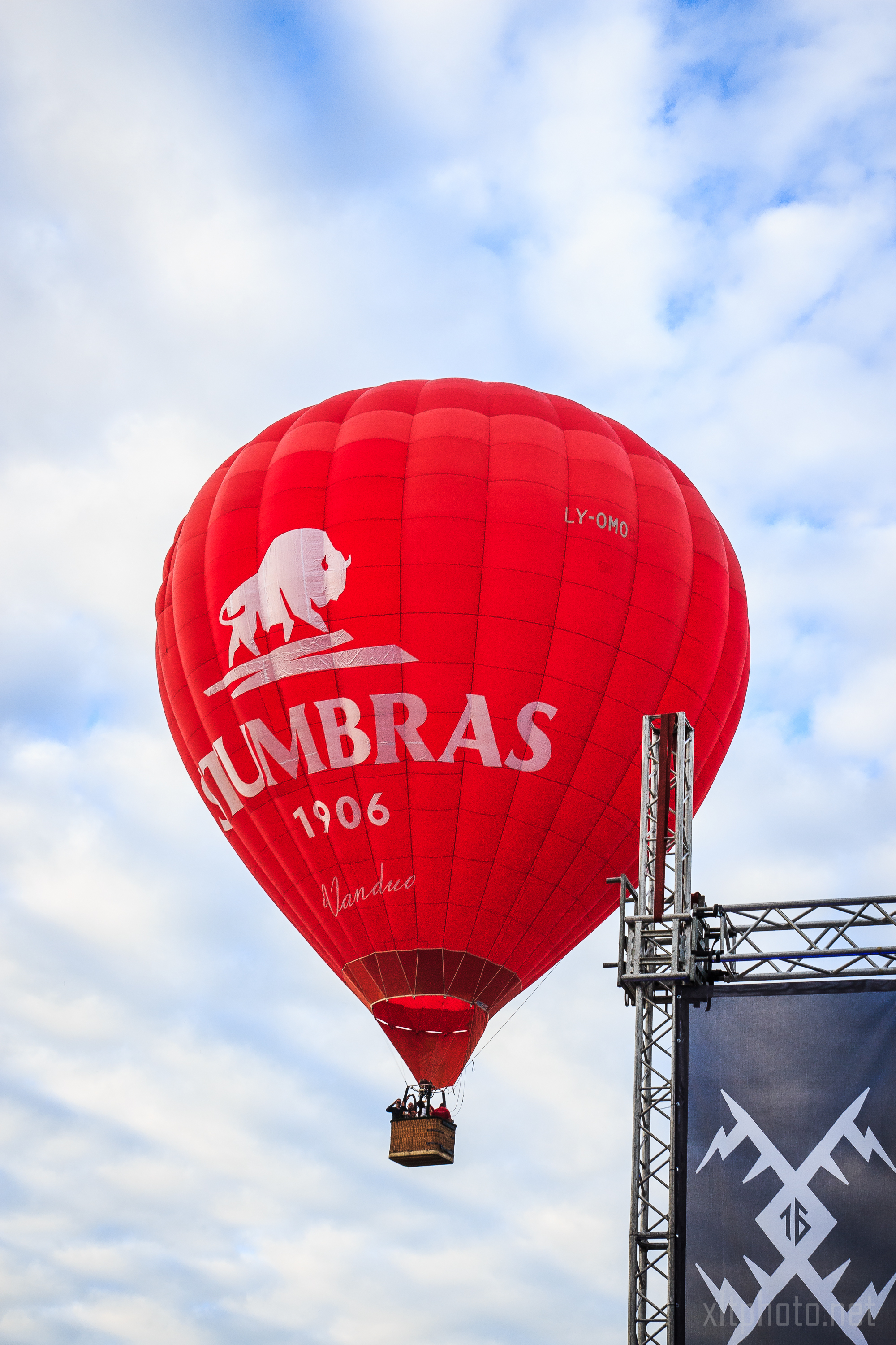 A red hot air balloon with the logo of "Stumbras 1906" and a white bison graphic rises against a blue sky with scattered clouds at the Kilkim Žaibu 2015 festival. The balloon's identification "LY-OMO" is visible. In the foreground is a metal scaffolding structure with a partial logo at the bottom right.