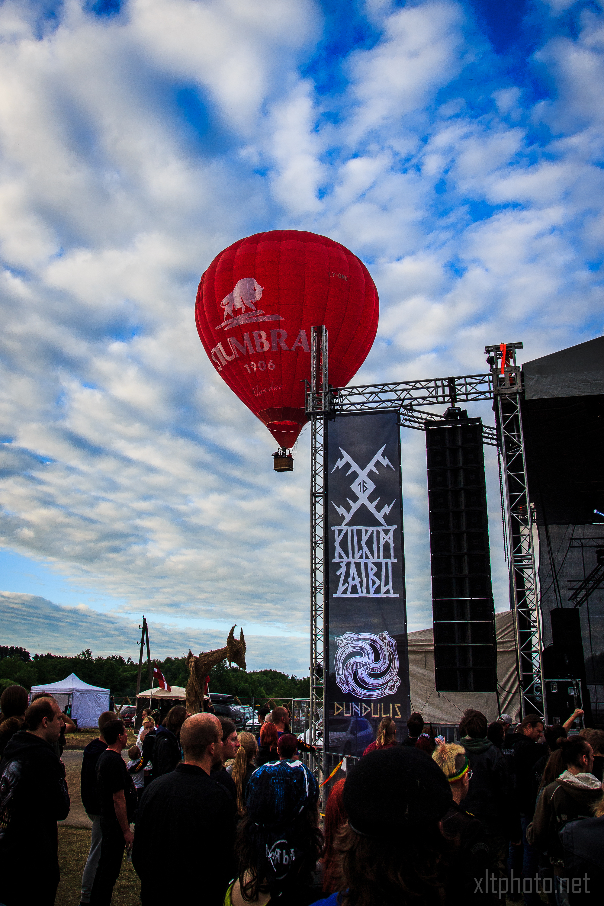 A bright red hot air balloon with white text floats above a crowd at the 2015 Kilkim Žaibu festival. Banners on the right bear logos and festival names. People on the ground observe the balloon amidst tents and structures under a partly cloudy blue sky. A mechanical dragon sculpture is visible in the background.