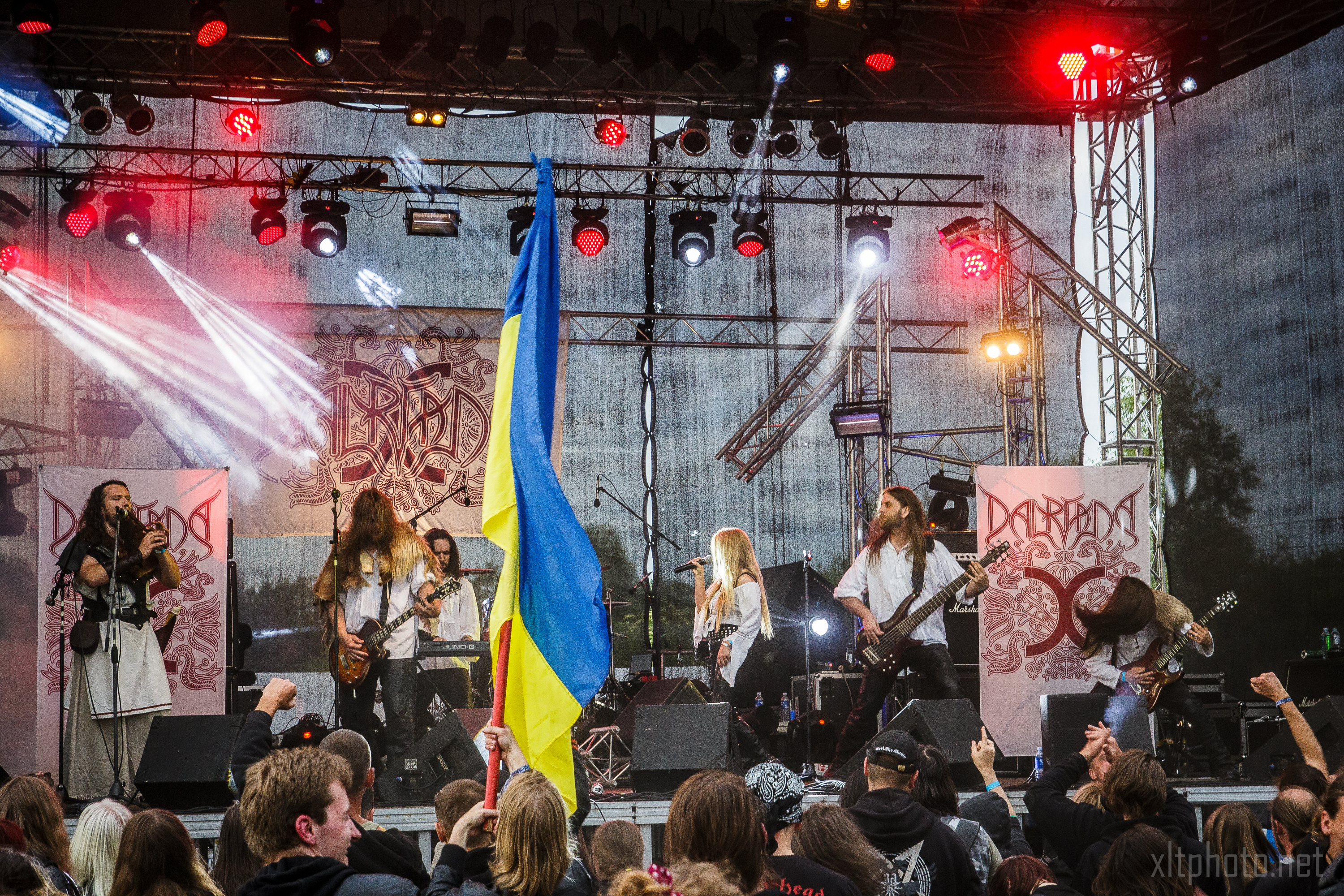 A band performs energetically on stage at an outdoor festival, with a large Ukrainian flag waved by an audience member in the foreground. The stage backdrop features the band's logo with "Kilkim Žaibu 2015" subtly included. The crowd is engaged, some raising their hands, all amid vibrant and intense lighting.