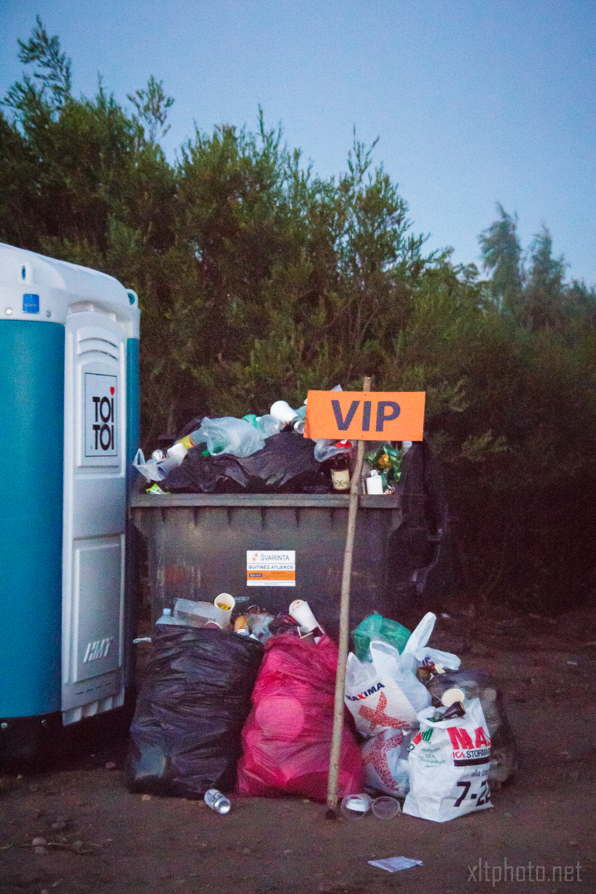A large outdoor garbage bin at the 2015 Kilkim Žaibu festival is overflowing with trash, surrounded by more bags on the ground. A makeshift "VIP" sign humorously adorns the bin, while a portable toilet peeks out on the left. Tall trees provide a natural backdrop to this quirky festival scene.