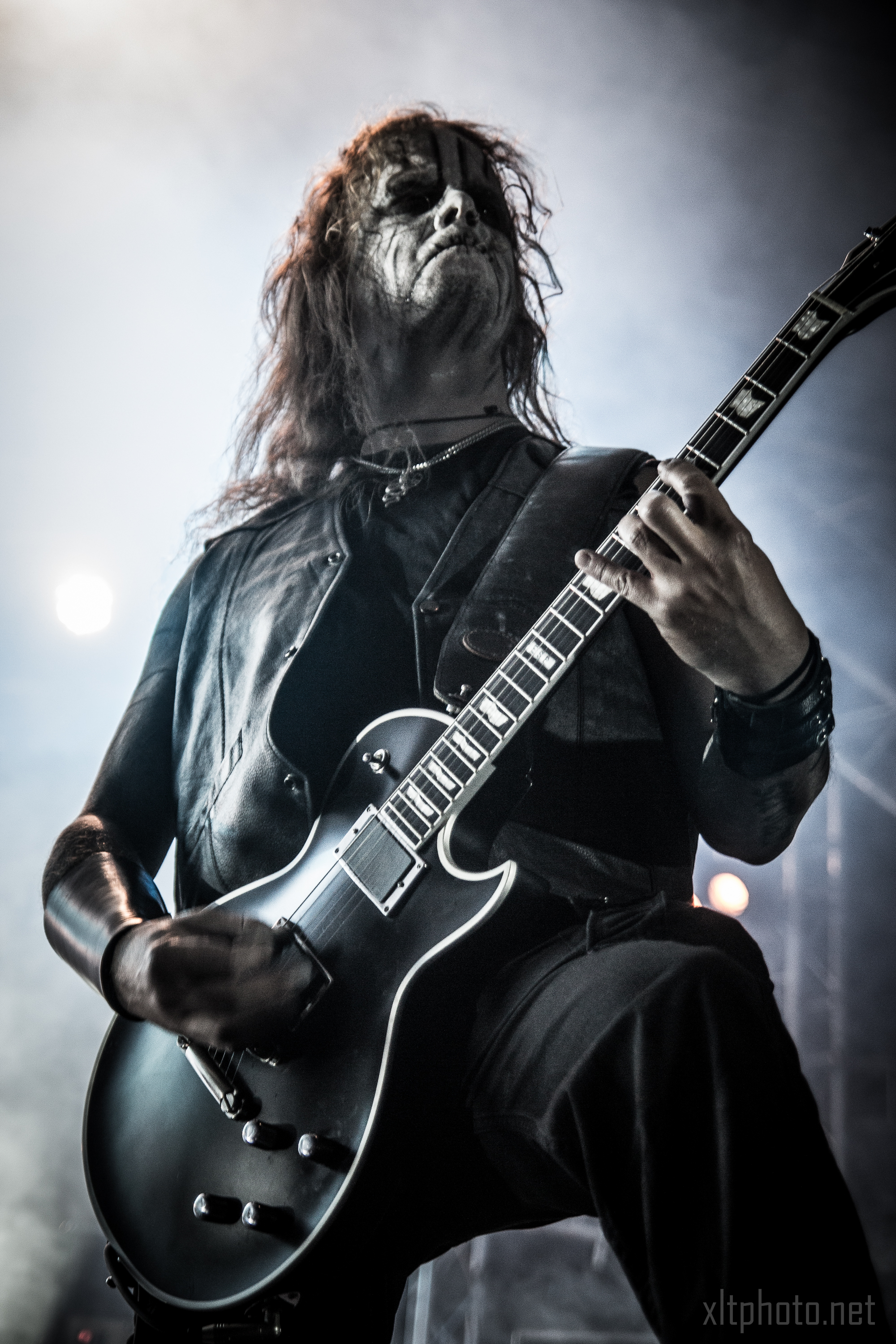 A musician with long hair and face paint, donning a black sleeveless leather vest, passionately plays an electric guitar on stage during the 2015 Kilkim Žaibu festival. The background is misty with stage lights creating an atmospheric effect. The photo is watermarked with "xltphoto.net" in the bottom right corner.
