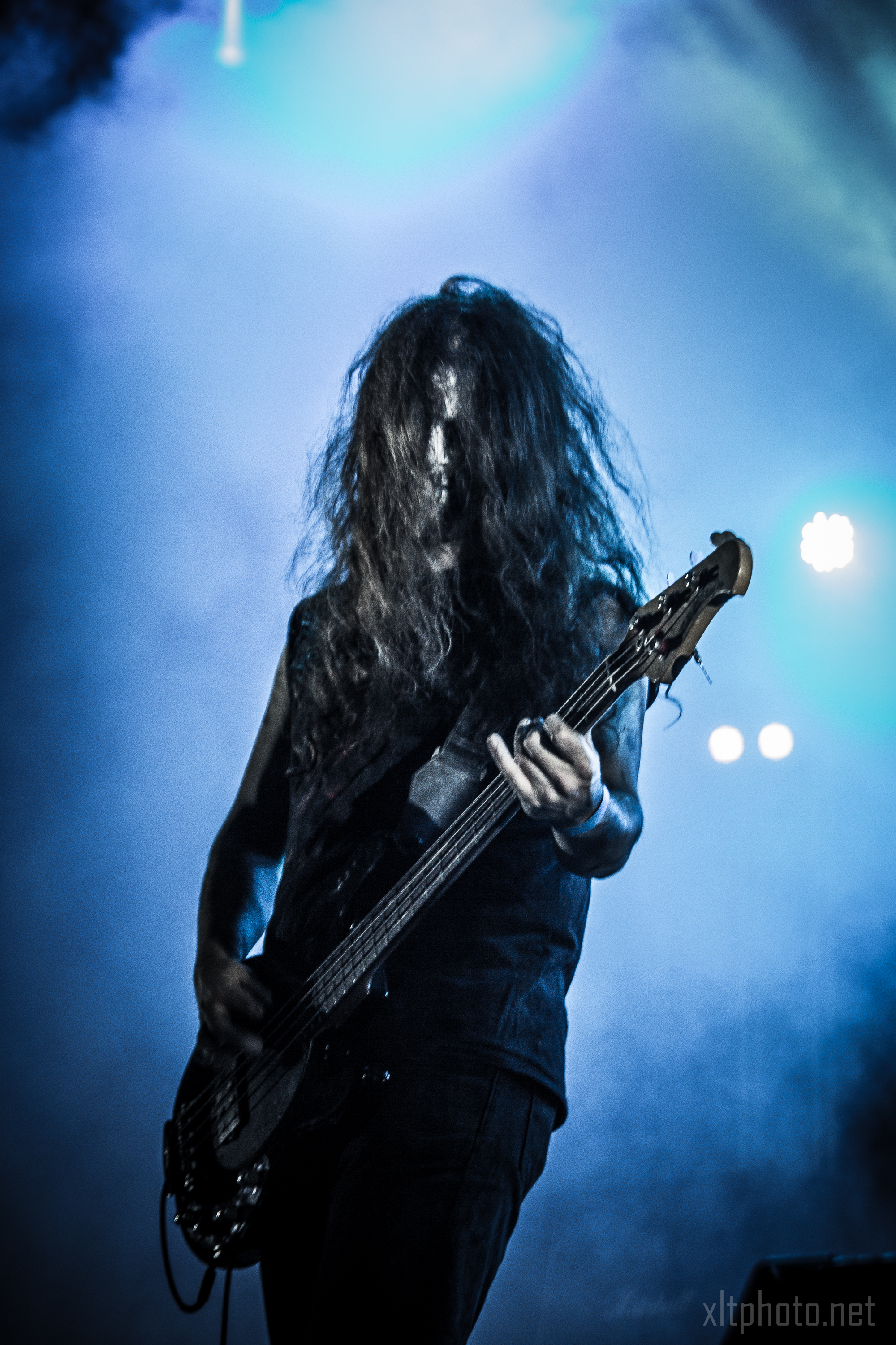 At the 2015 Kilkim Žaibu festival, a musician with long, wavy hair performs on stage. Their hair obscures their face as they play a bass guitar. The background is filled with blue and black lighting, creating a dramatic and intense atmosphere, while their dark outfit enhances the moody setting.