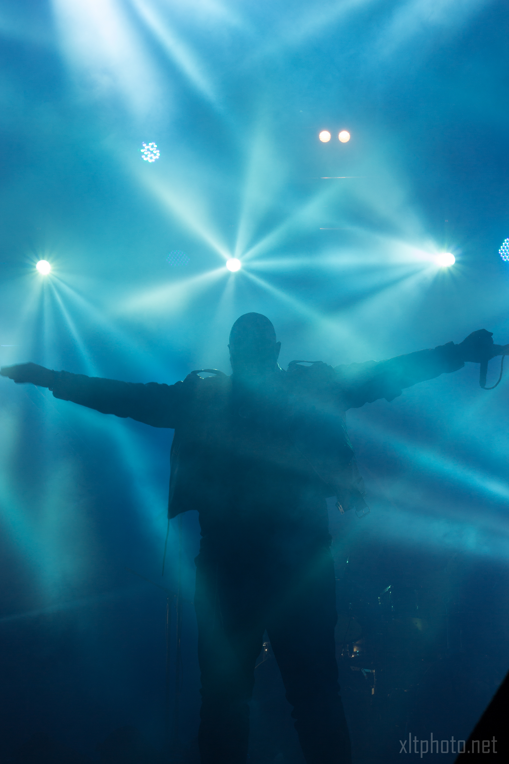 A silhouetted figure stands with outstretched arms amidst a blue, hazy environment. Bright stage lights beam from behind, illuminating the background with starburst effects. The person, holding a microphone, adds an ethereal and dramatic touch reminiscent of Kilkim Žaibu festival 2015.