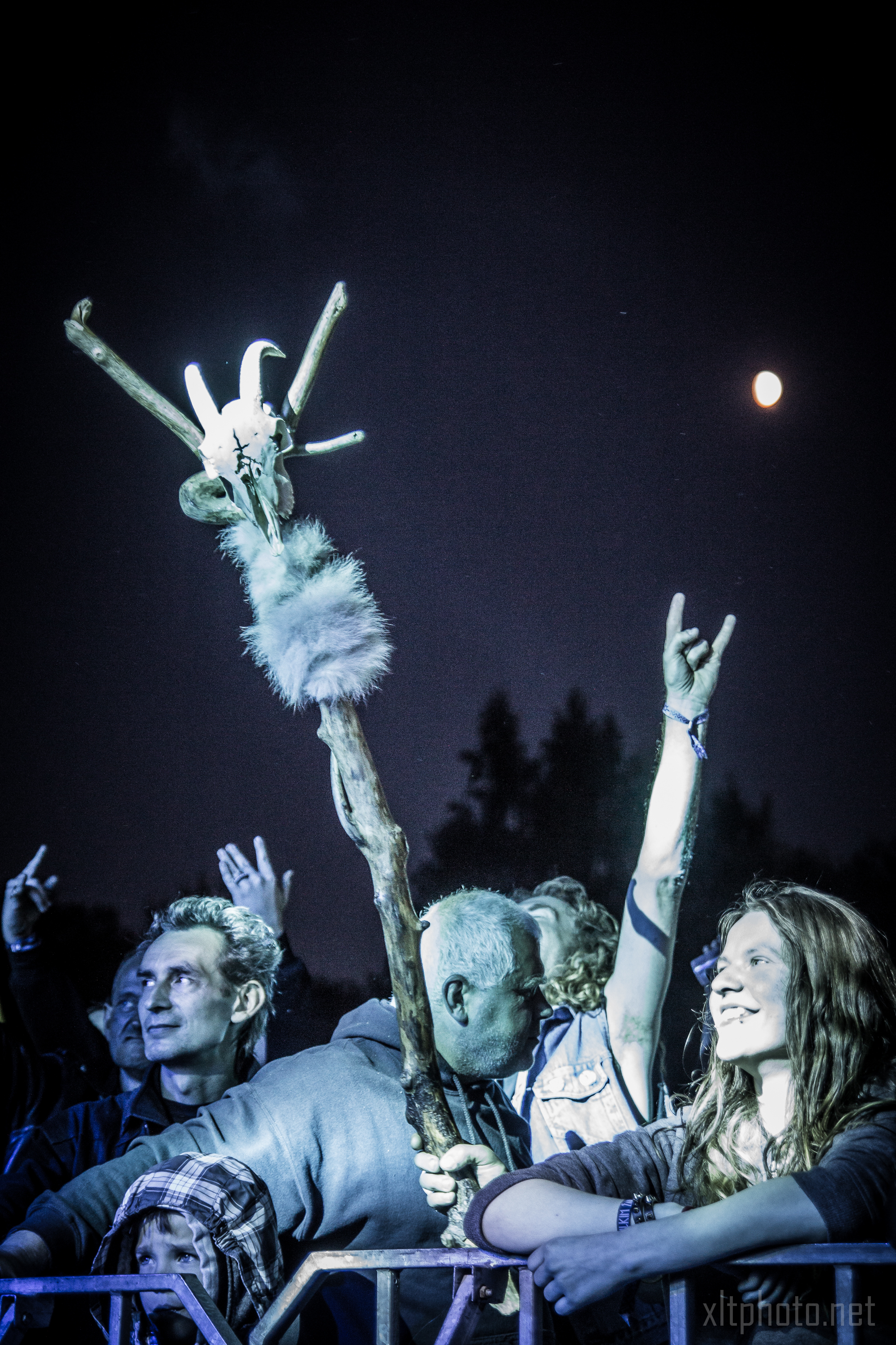 A crowd at the 2015 Kilkim Žaibu festival, with a full moon in the background. People are raising their hands, some making rock and roll signs. A prominently raised wooden staff adorned with feathers, fur, and a skull stands out in the crowd. The atmosphere is energetic and festive.