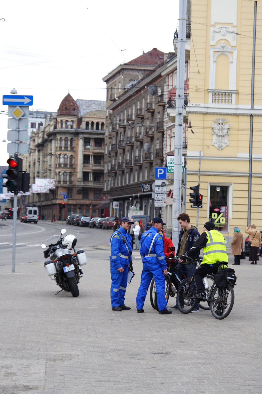 A street scene from 2010 with three people in blue uniforms and one in a yellow vest standing next to two bicycles. A police motorcycle is parked nearby. They're conversing on a city sidewalk amidst ornate historic buildings, possibly during a Kritiska masa event, with a traffic light and distant pedestrians in the background.