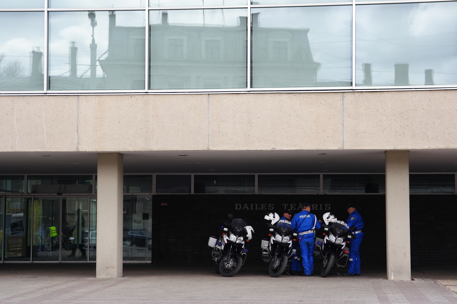 Three police officers, dressed in blue uniforms, are standing beside three police motorcycles under a concrete overhang. The building behind them features large windows on the upper floor and the words "DAILES TEATRIS" on the dark wall beneath the overhang. This scene evokes memories of Kritiska masa 2010.