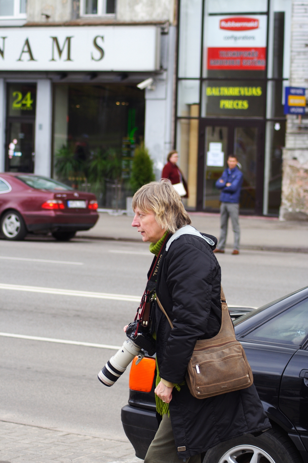 A person with short, light hair is walking on a city sidewalk, carrying a camera with a large lens. They are wearing a dark jacket and an olive scarf, and have a brown shoulder bag. In the background, cars are parked along the street, and people are standing in front of shopfronts during Kritiska masa 2010.