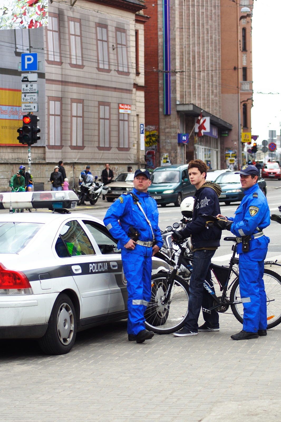 Two police officers in blue uniforms stand near a police car and a young man with a bicycle on a city street, reminiscent of the cycling event Kritiska masa in 2010. The conversation appears casual. Behind them, the cityscape features buildings, traffic lights, and pedestrians, suggesting routine urban activity.