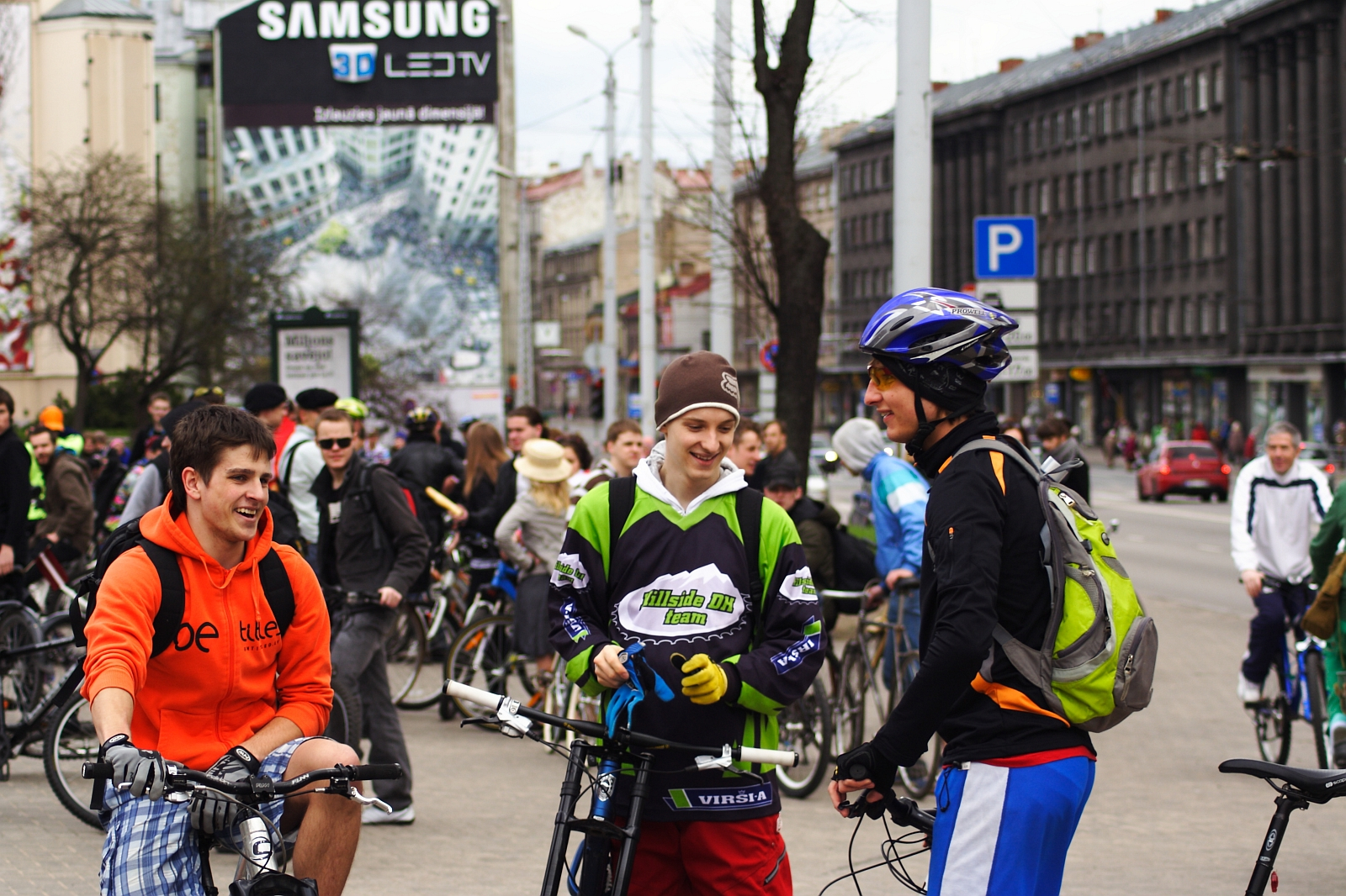 A group of people are gathered in a bustling city street with bicycles, reminiscent of Kritiska masa events around 2010. Three men in the foreground, smiling and chatting in casual and sporty attire with helmets and backpacks, stand out. The background features tall buildings, a "Samsung" advertisement, and more cyclists.