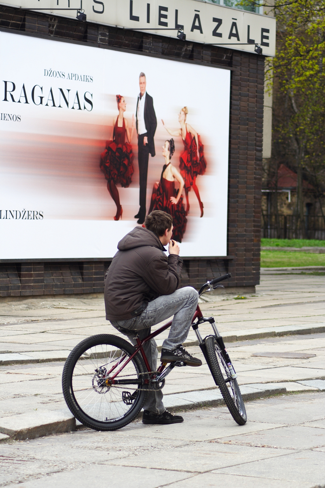 A person in a brown jacket and gray jeans is sitting on a bicycle on a paved walkway, reminiscent of the 2010 Kritiska masa. They appear to be eating an ice cream cone. Behind them, there is a large advertisement featuring a man in formal attire and women in red and black flamenco dresses. The sign has text in a non-English language.