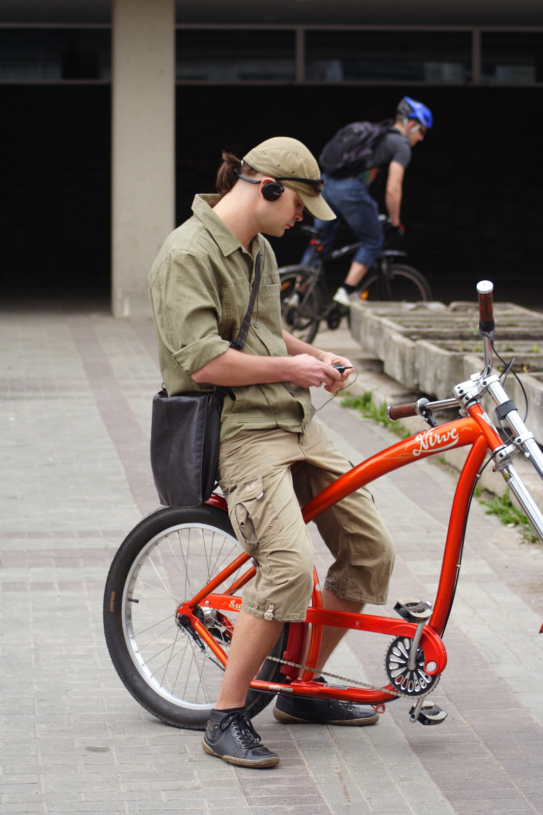A young man in khaki shorts and a matching shirt sits on a bright orange bicycle. He wears a black cap with headphones, and a black shoulder bag across his body while looking at a smartphone. In the background, another cyclist with a blue helmet rides by during the 2010 Kritiska masa event in an outdoor urban area.